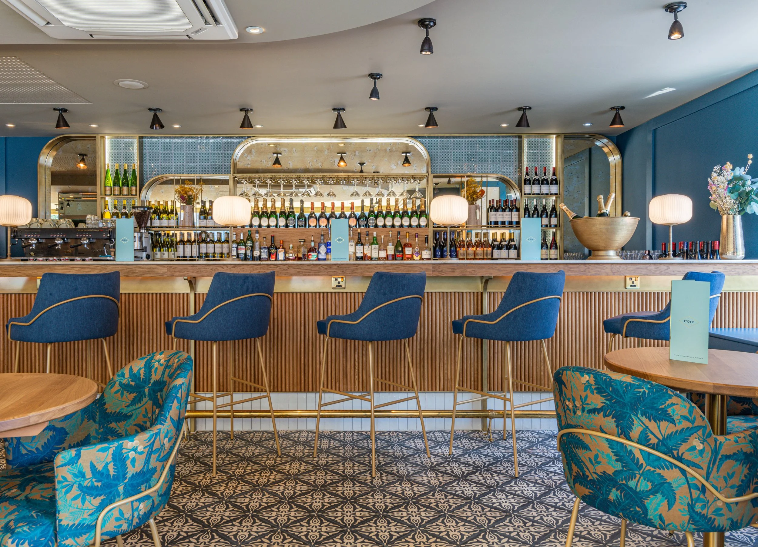 Stylish modern bar interior with blue chairs and patterned flooring, featuring a well-stocked bar with liquor bottles and wine glasses. Round tables and floral arrangements add to the decor.