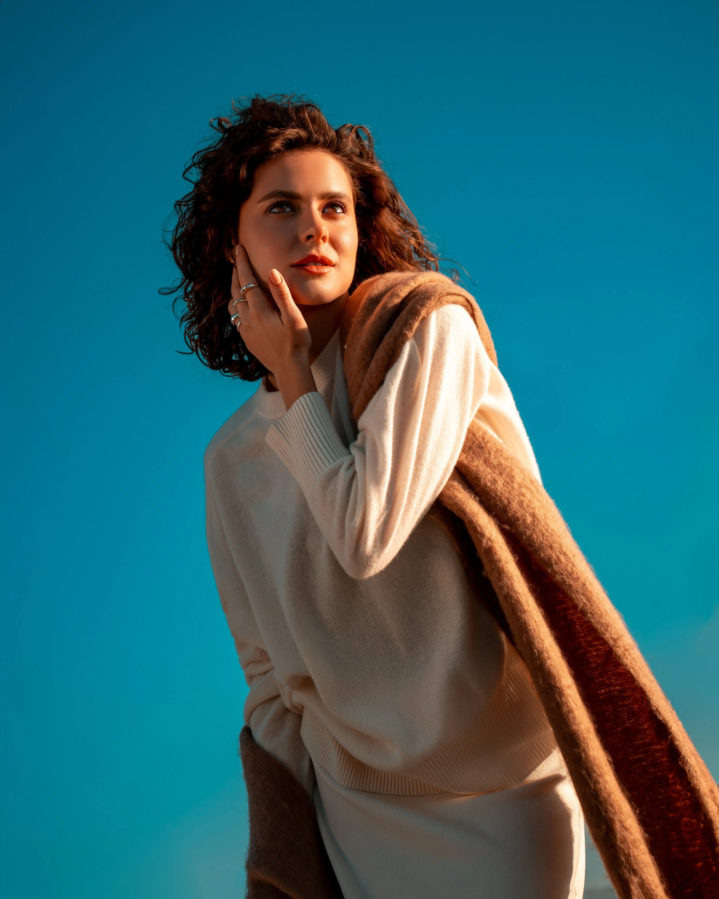 Woman with curly hair wearing a white sweater and brown scarf, gazing thoughtfully, with a clear blue sky background.
