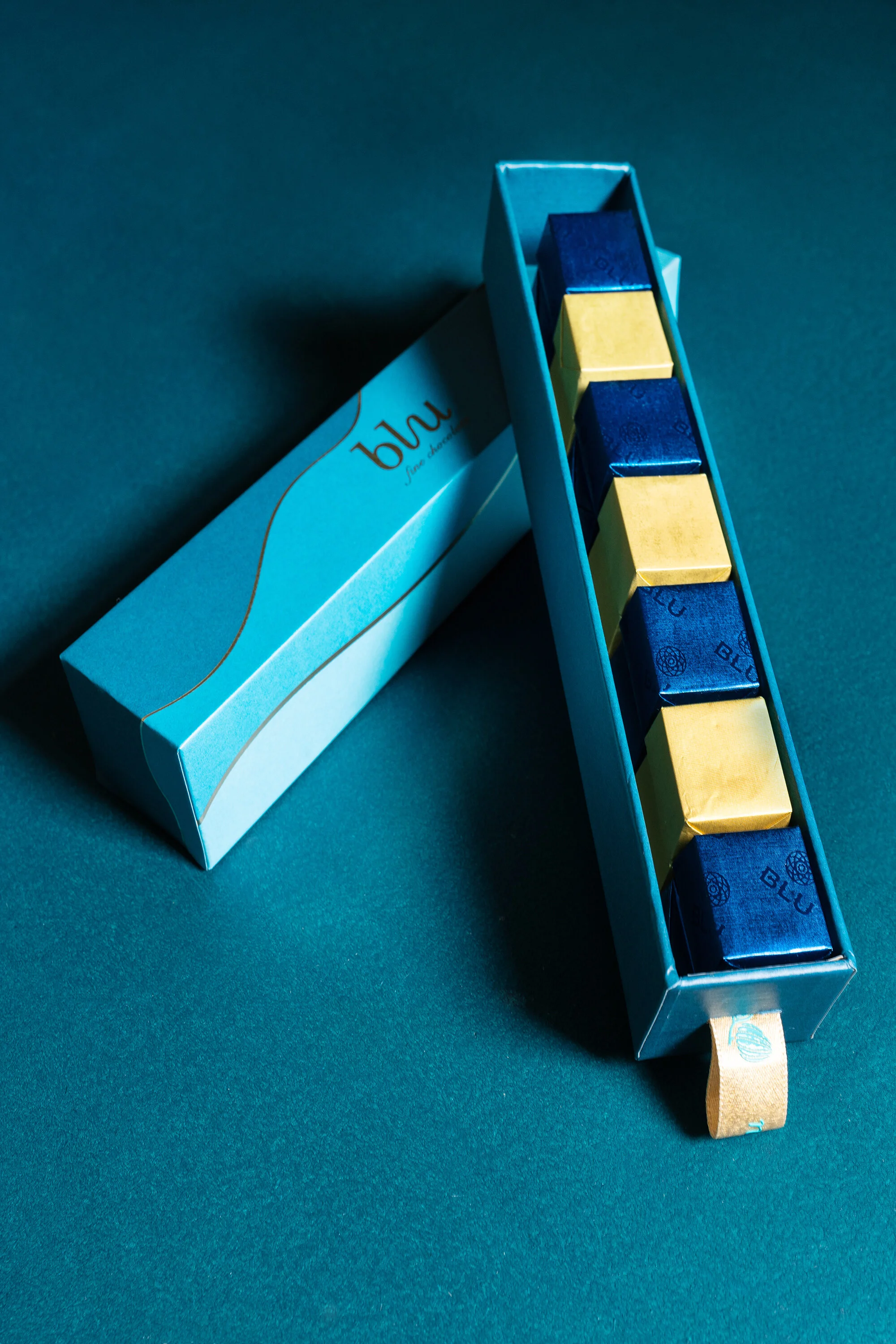 Box of individually wrapped chocolates in blue and gold packaging on a teal background.