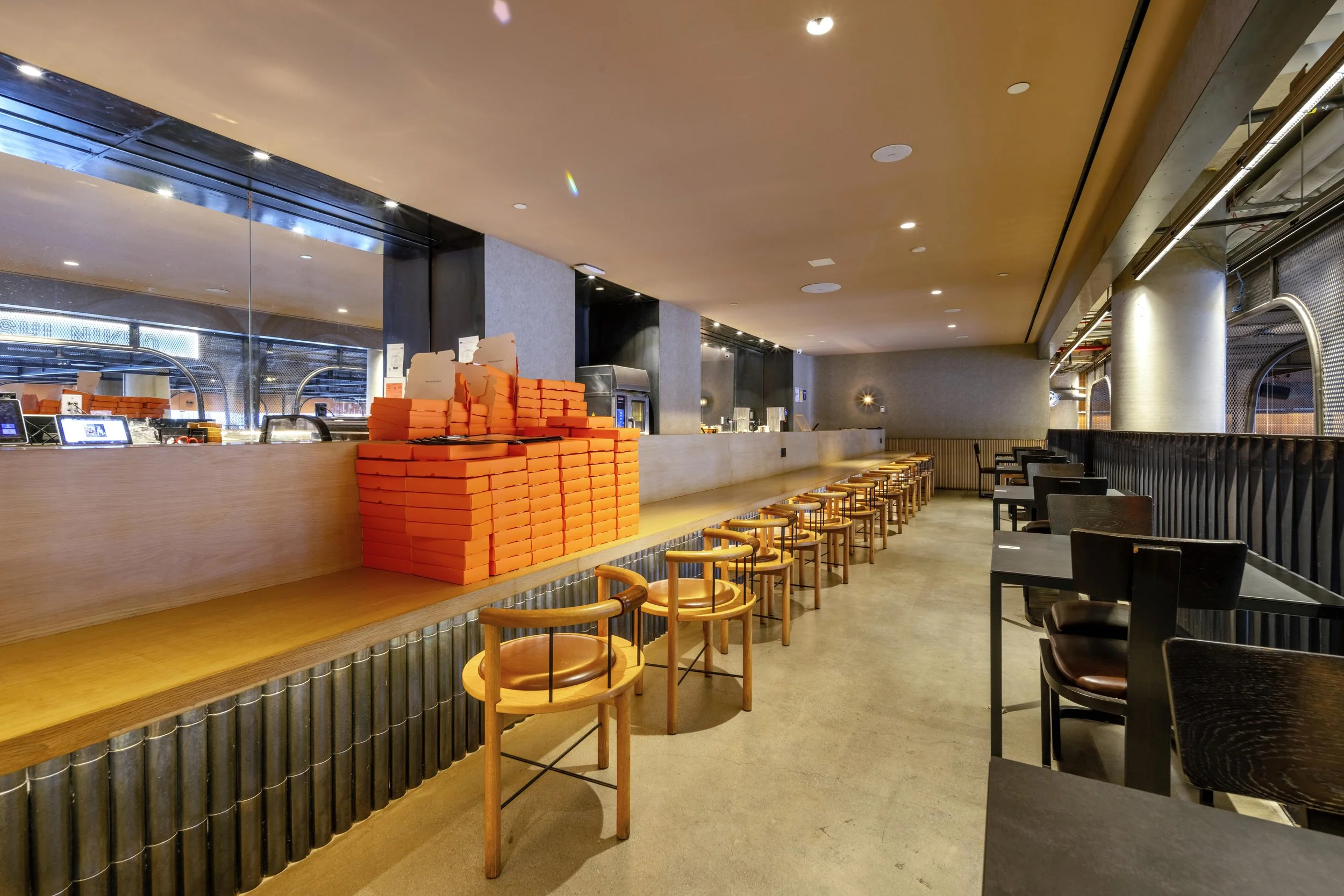 Modern café interior with wooden chairs and tables, orange pizza boxes stacked on the counter, and ambient lighting.