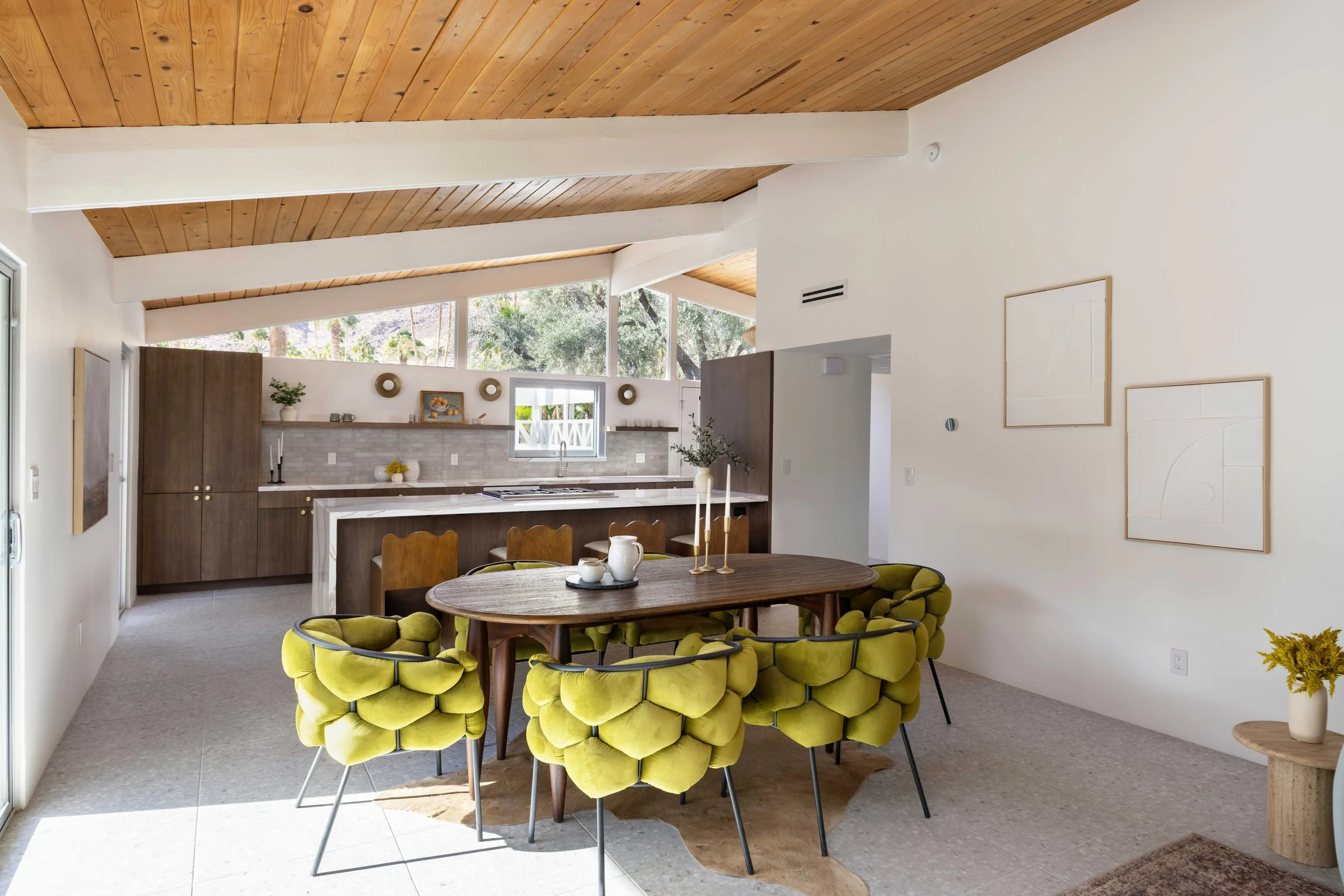 Modern open-plan kitchen and dining area with wood ceiling, green cushioned chairs around a wooden dining table, and a kitchen island in the background.