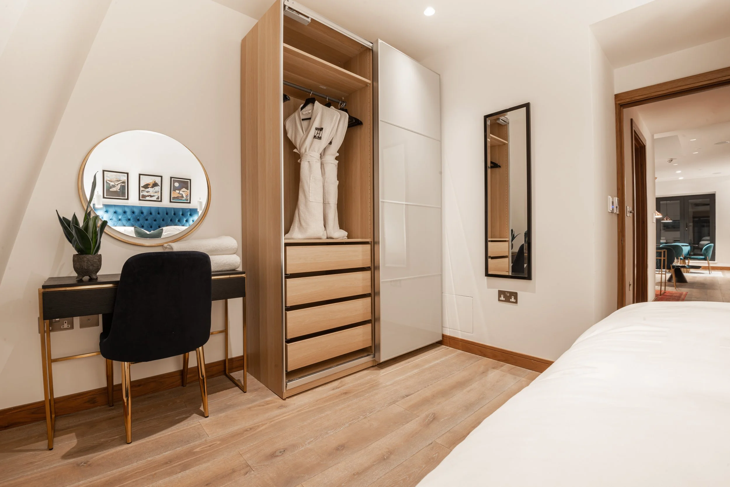 Modern bedroom with wardrobe, desk with mirror, black chair, and indoor plant; hardwood floors and open wooden door leading to a living area.