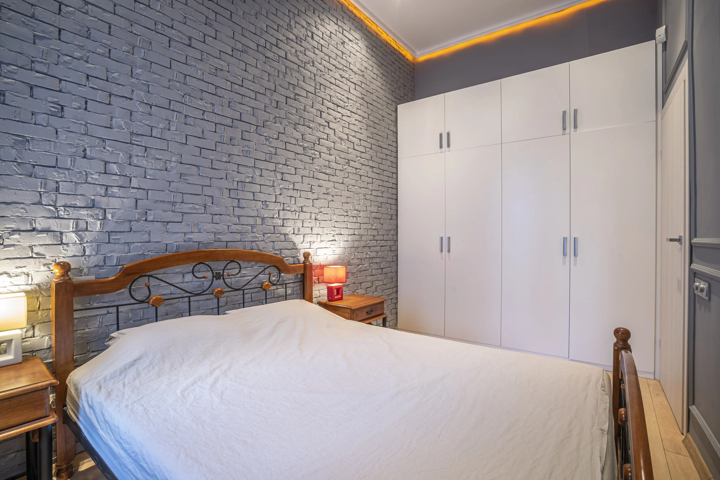 Cozy bedroom with a blue brick accent wall, wooden bed frame, white bedding, white cupboards, wood side tables, and warm lighting.