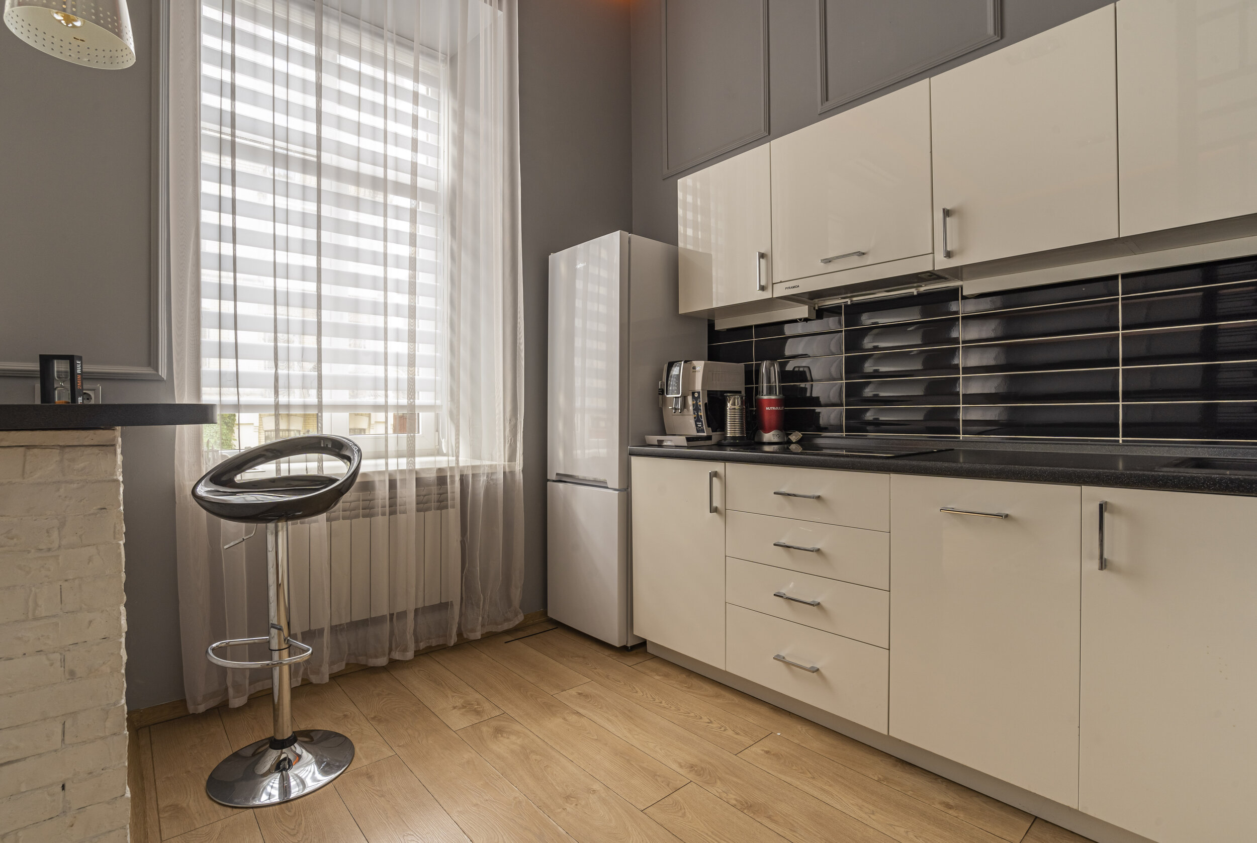 Modern kitchen interior with white cabinets, black backsplash, and wooden flooring. Features a refrigerator, coffee machine, high stool, and striped curtains on a large window.