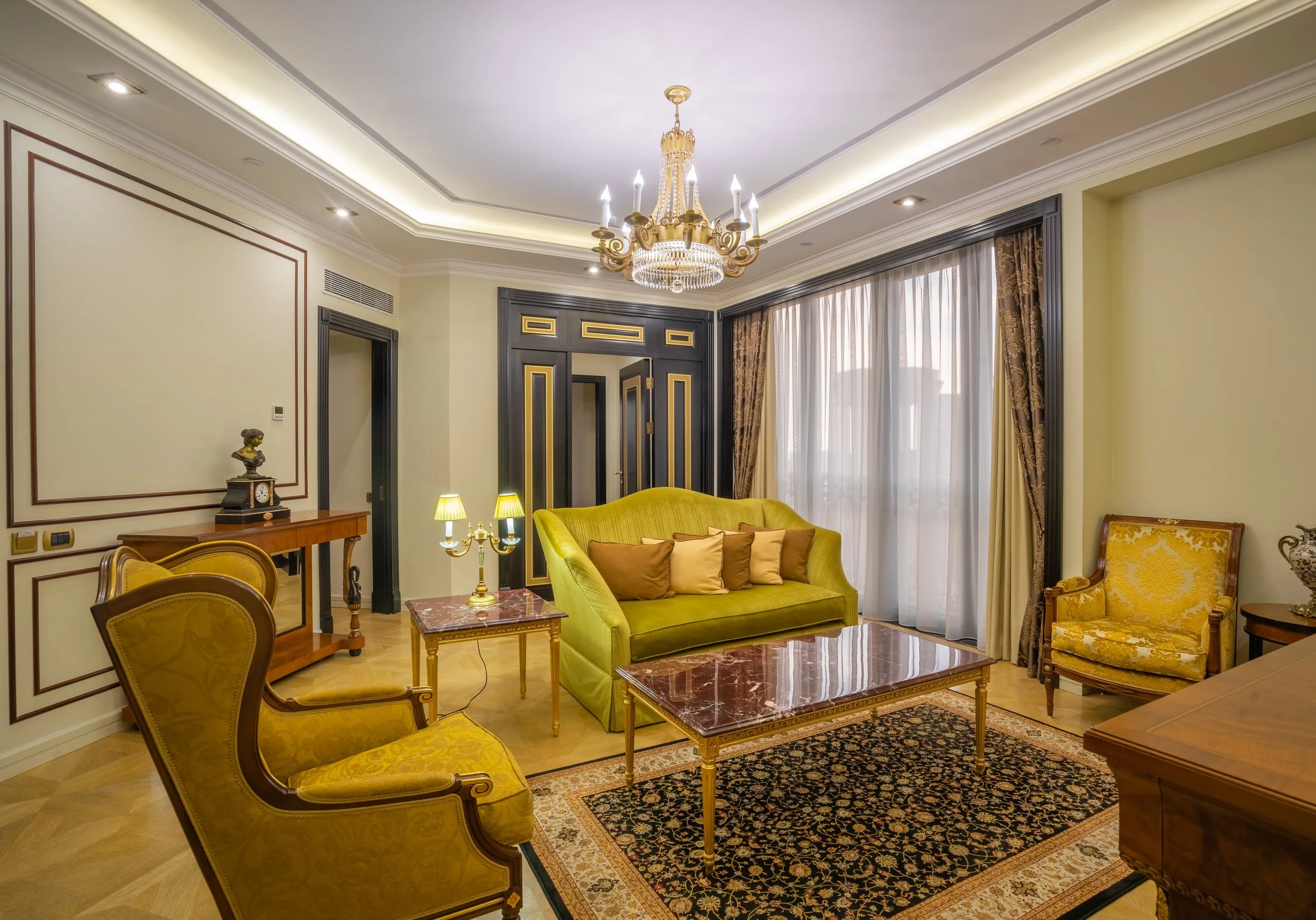 Elegant living room with a chandelier, yellow armchairs, green sofa, marble-top tables, and patterned rug.