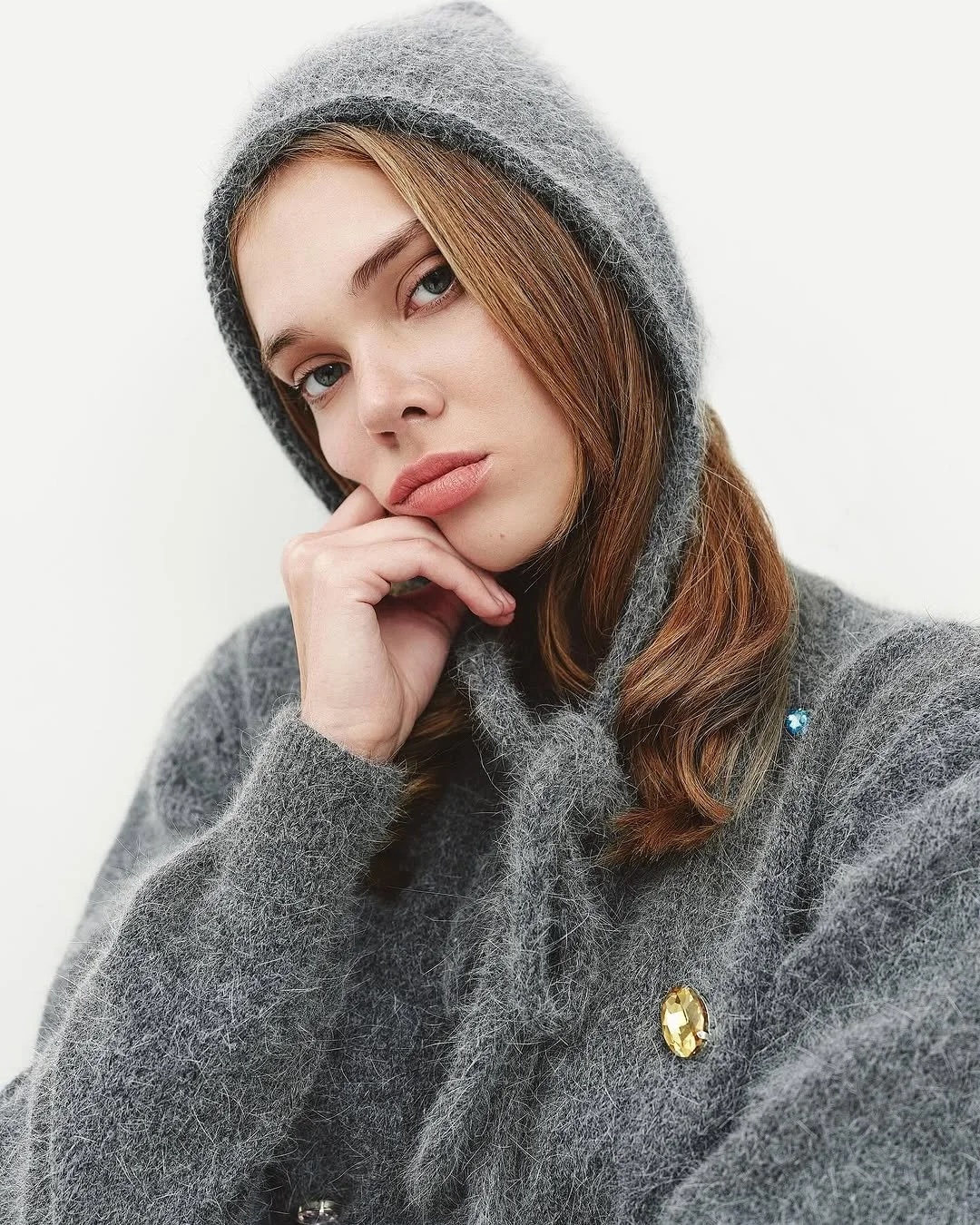Person wearing a gray fuzzy hooded sweater with decorative gems, resting their chin on their hand, looking at the camera.