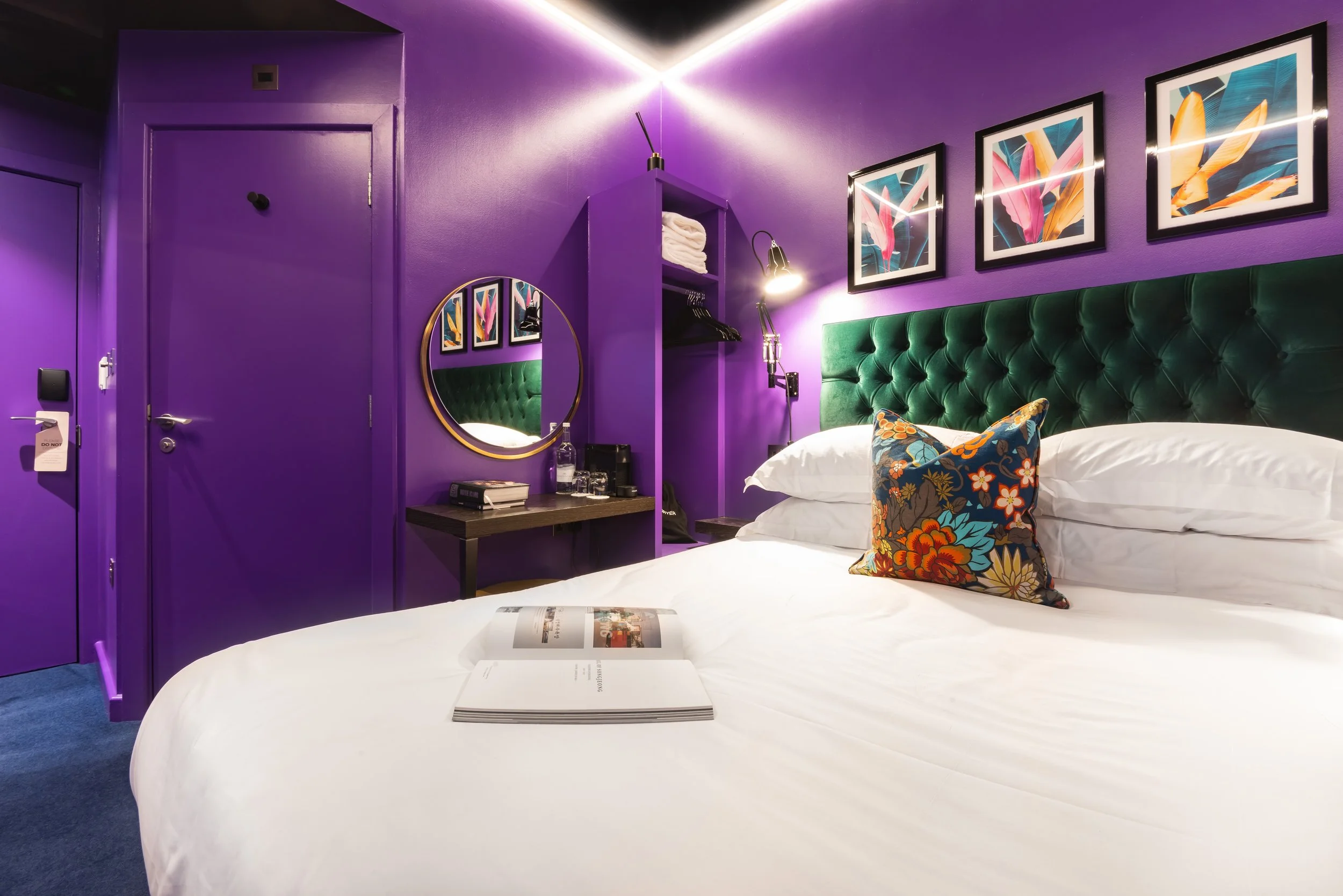 Modern hotel room with vibrant purple walls, green tufted headboard, and colorful decor. Bed with white linens and floral pillow, artwork on walls, a mirror, bedside table with books and water, and closet with shelves.