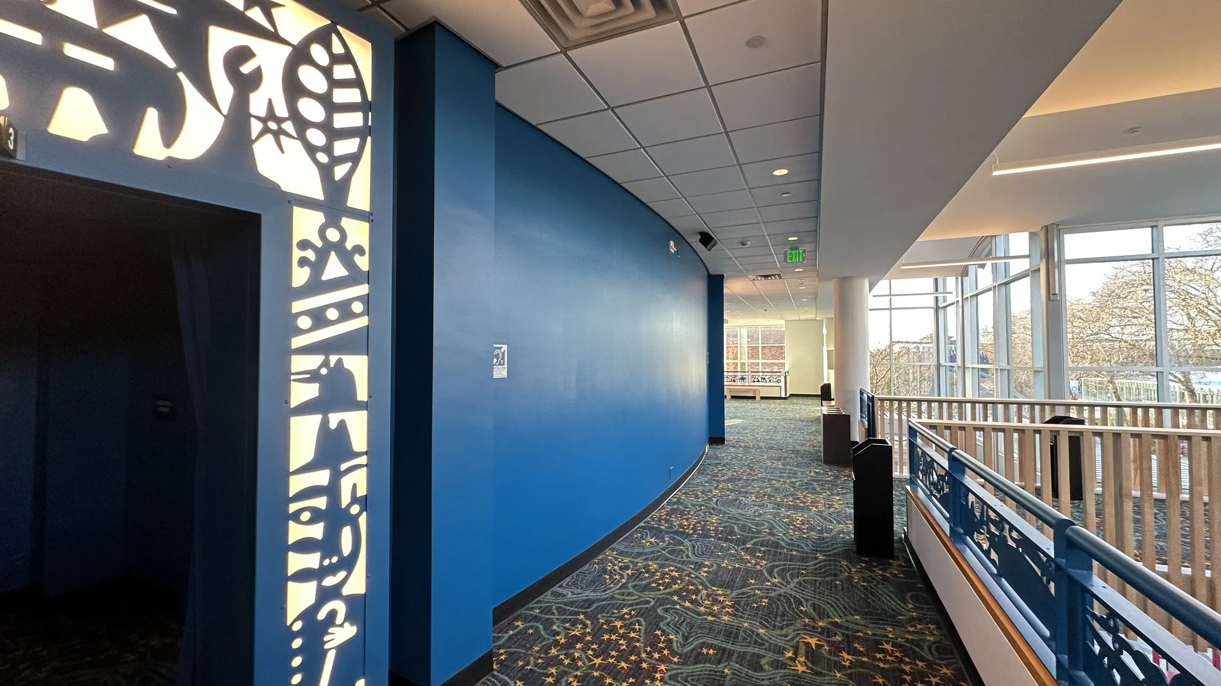 Empty hallway with blue walls, patterned carpet, large glass windows, and wooden railing, in a modern building.