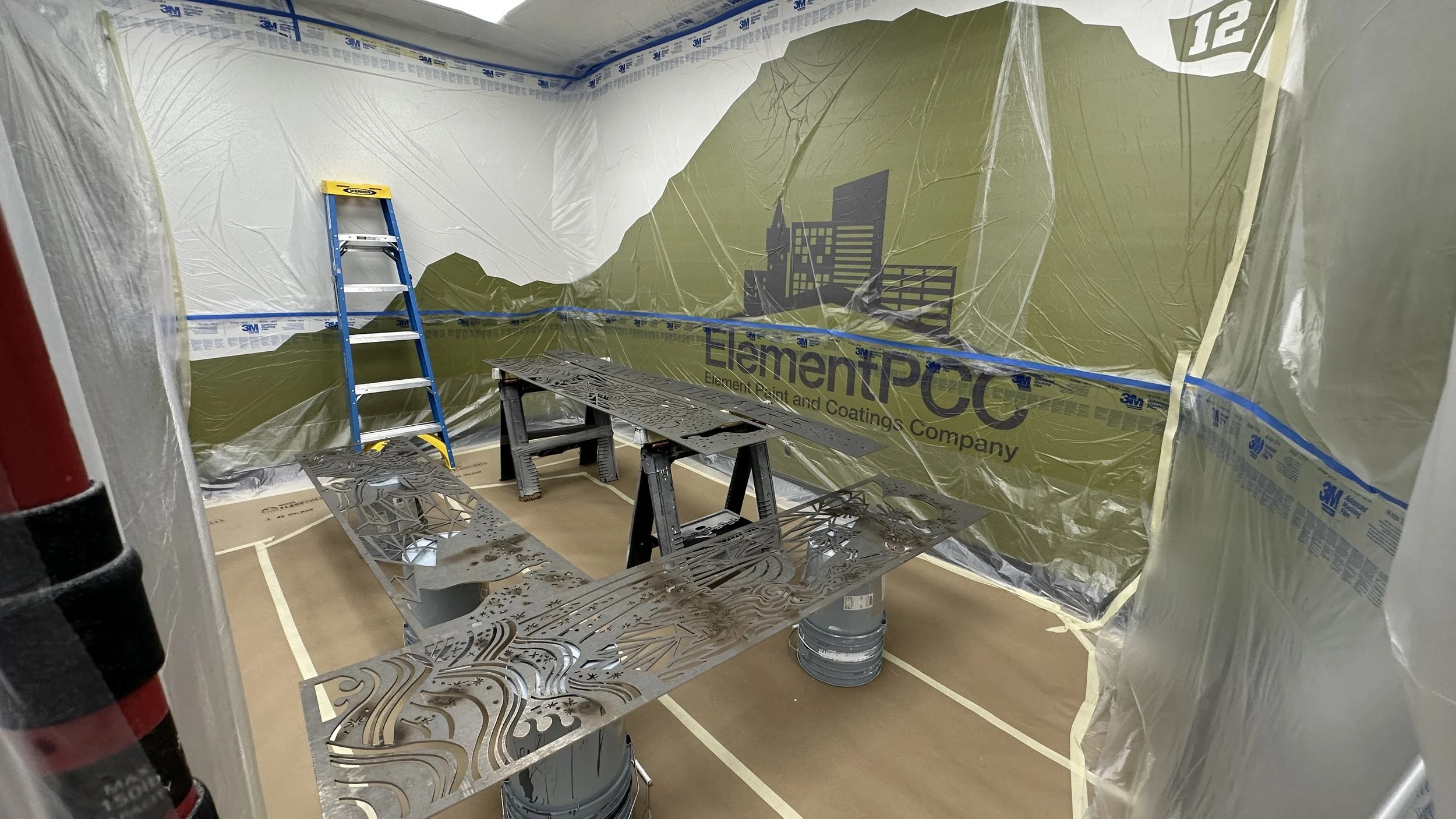 A room under construction with protective plastic sheeting on the walls, a blue ladder, and metal cutouts on top of paint cans.