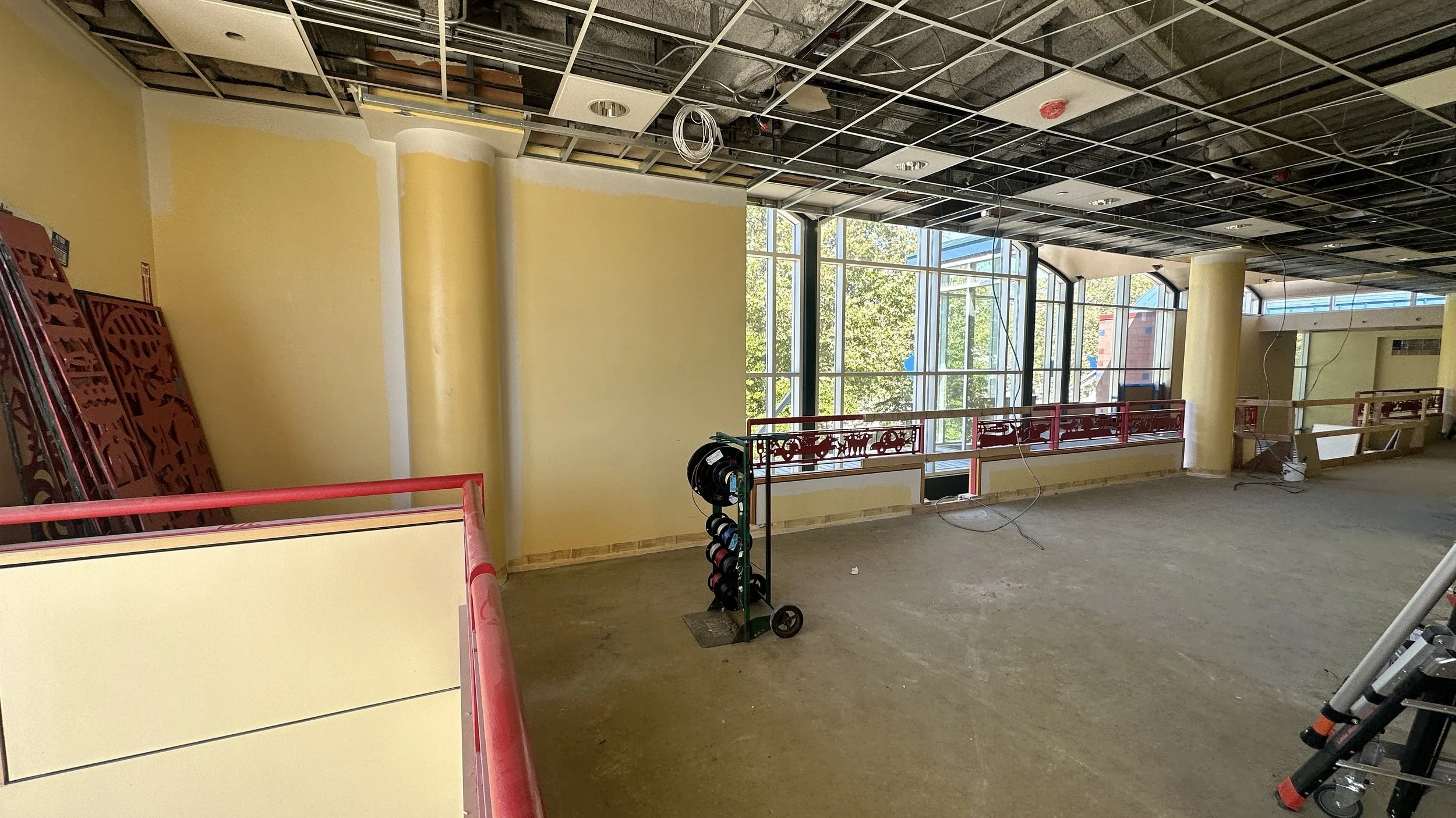 Interior of a building under construction with yellow walls, large windows, and exposed ceiling with visible wiring and piping. Construction tools are present, including a hose reel and scaffolding, with a red railing around the edge of the upper level.