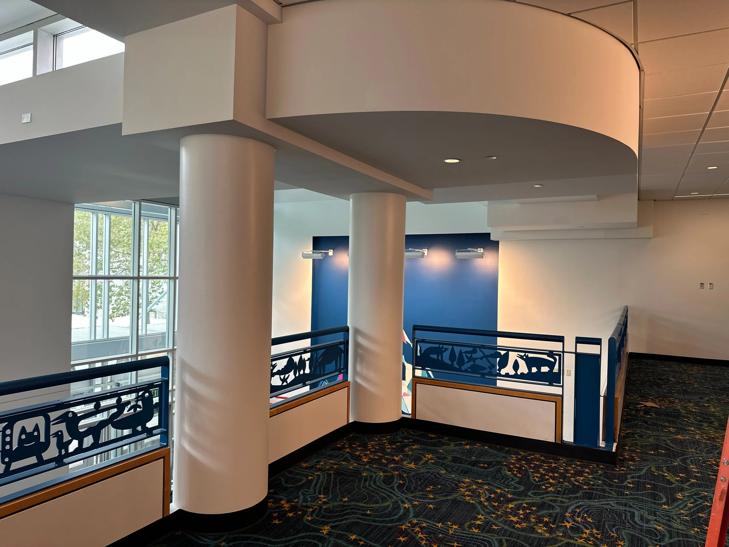 Interior of a modern building with white walls, large windows, and a staircase with decorative blue railing featuring various playful silhouettes. The floor is carpeted with a dark, patterned design.