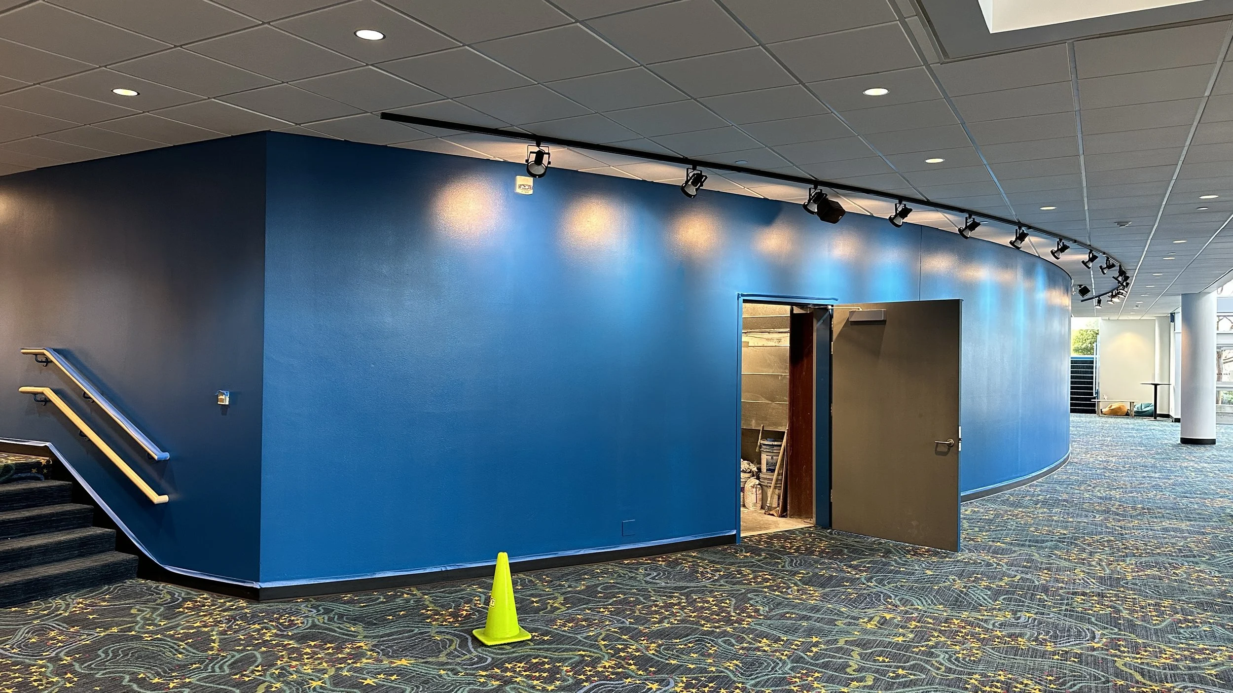 Interior view of a modern building with a curved blue wall, staircase on the left, partially open door revealing a storage room, and a colorful patterned carpet. Overhead lighting and a row of ceiling spotlights are visible.