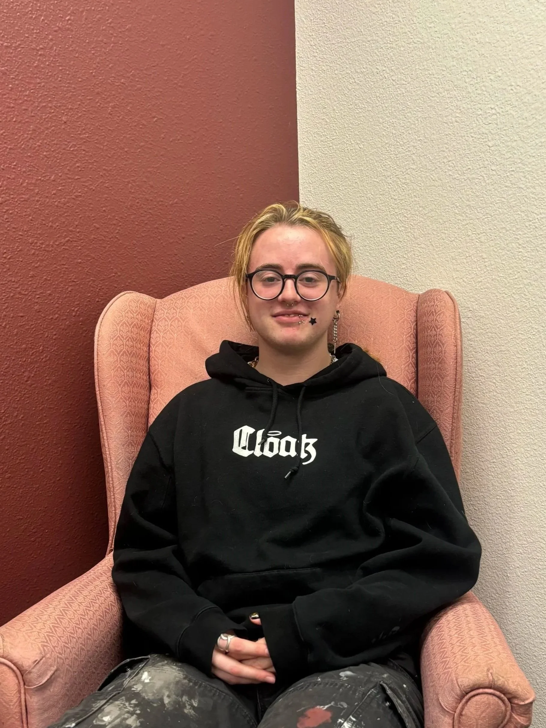 A person with blonde and orange hair, wearing glasses, a plaid shirt, and a septum piercing, sitting indoors with a window in the background showing a parked vehicle outside.
