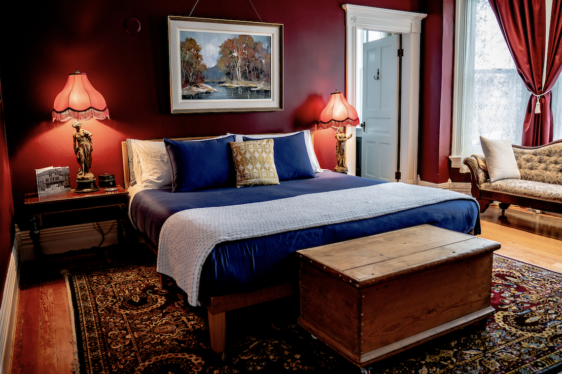 A bedroom with dark red walls, a double bed with blue bedding and pillows, two bedside lamps with pink lampshades, a wooden trunk at the foot of the bed, a large painting of a river and trees above the bed, a window with white trim and red curtains, and a vintage sofa with a pillow near the window.