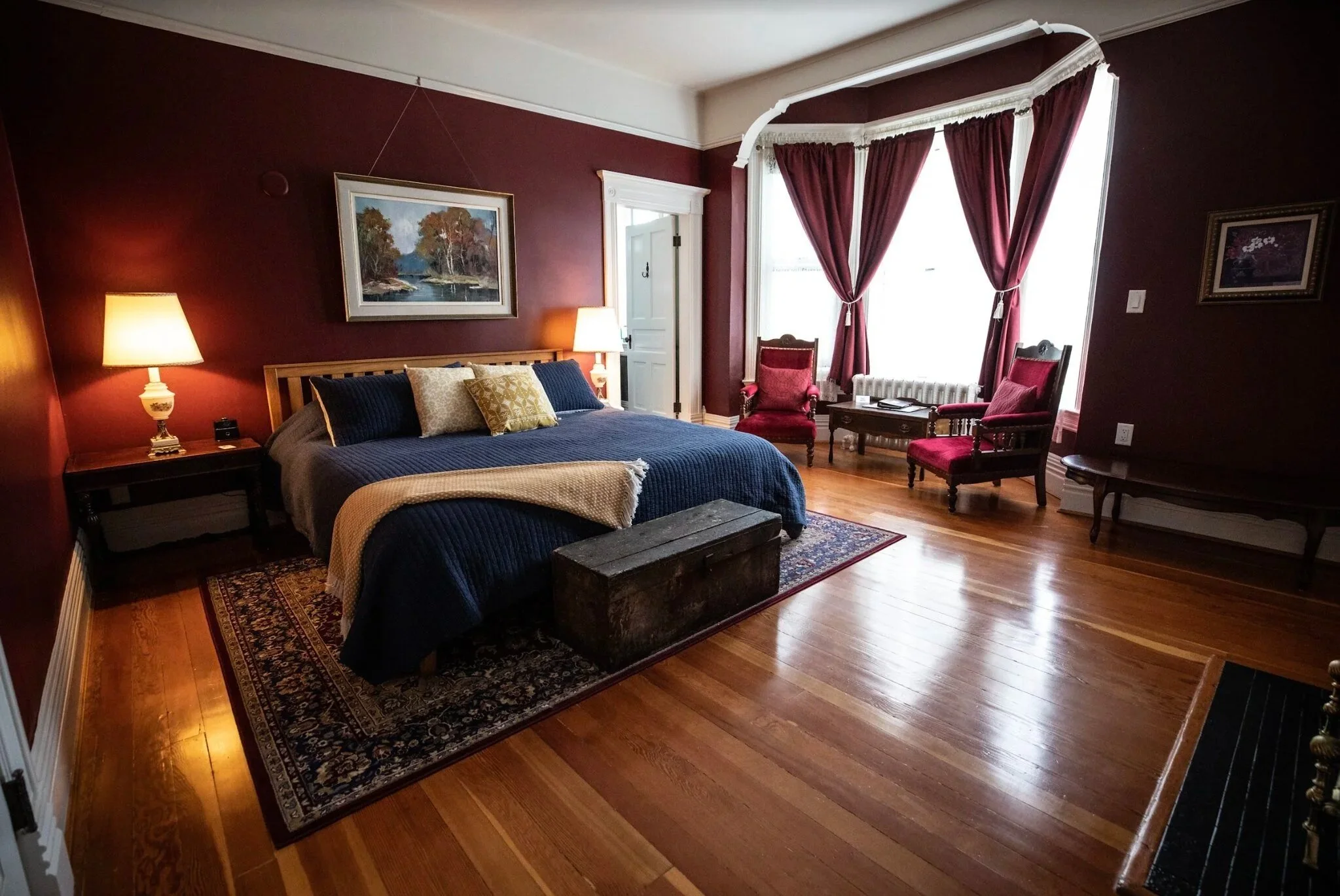 Guest Room at The Wild Turkey Inn. A bedroom with dark red walls, a large bed with a navy blue quilt, and decorative pillows. The room has hardwood floors and framed artwork on the walls.