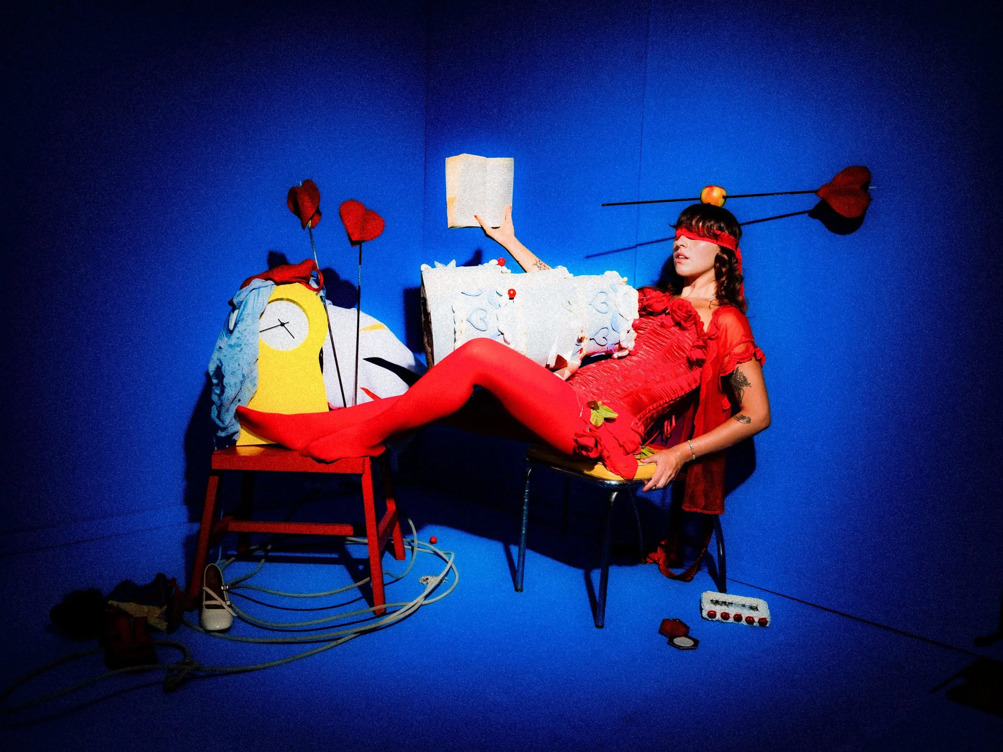 A woman in a red dress and blindfold, reclining on a chair with blue walls, surrounded by props including a clock, red hearts on sticks, and an arrow through a heart on her head.