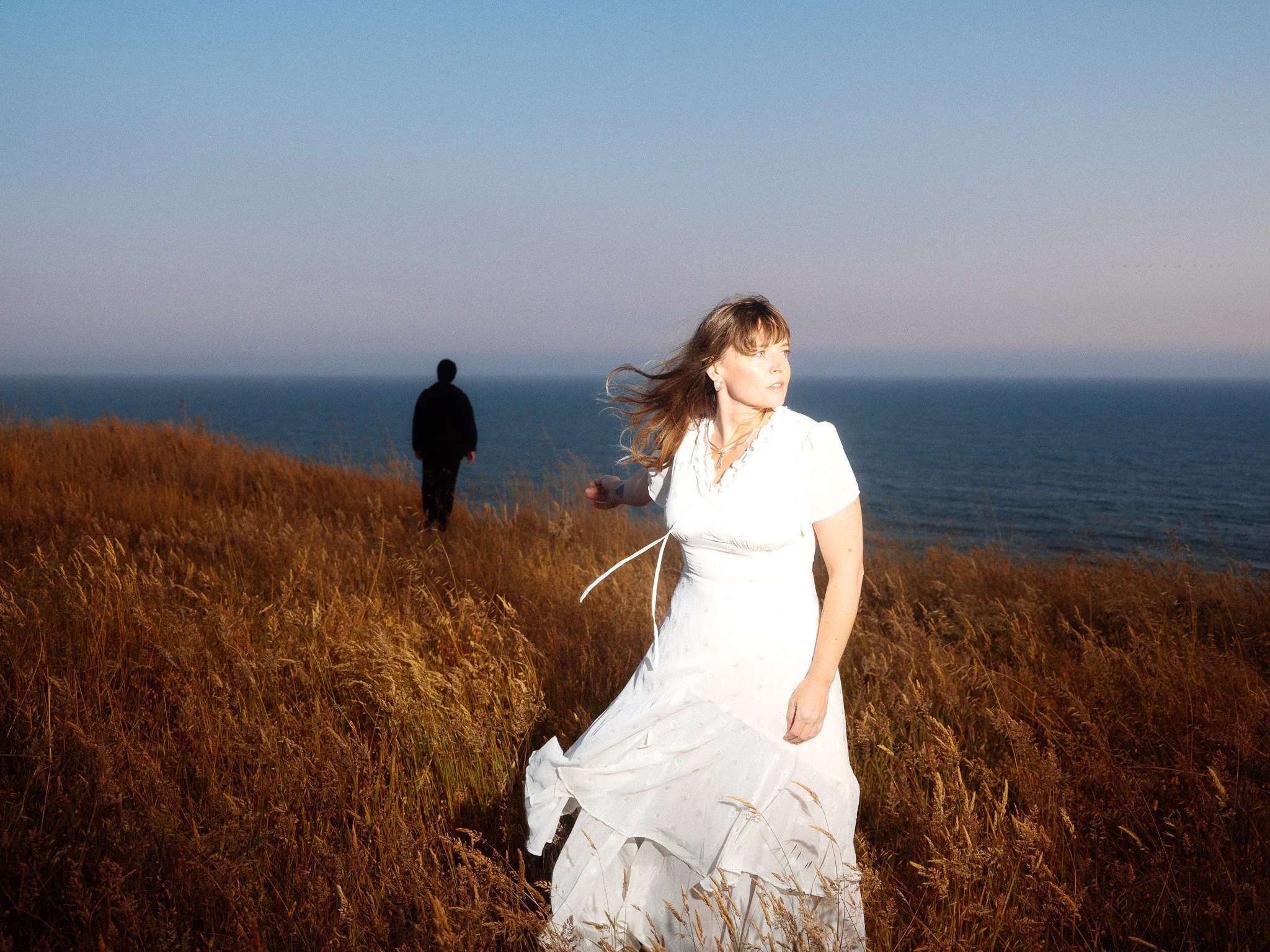 A woman in a white dress standing in grassy field near the coast, with a man walking away in the background, ocean and sky in the distance.