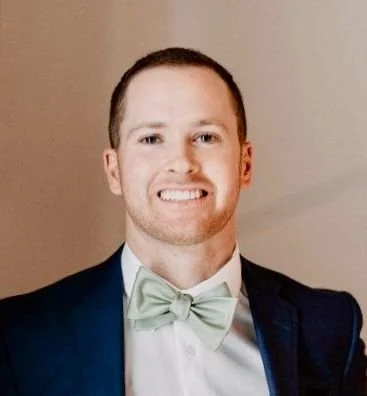 A man in a tuxedo with a mint green bow tie, smiling against a neutral background.