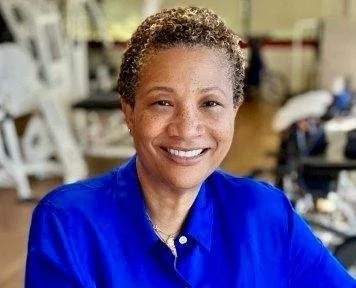 A woman with short curly hair smiling, wearing a blue shirt, in a workshop or craft room.
