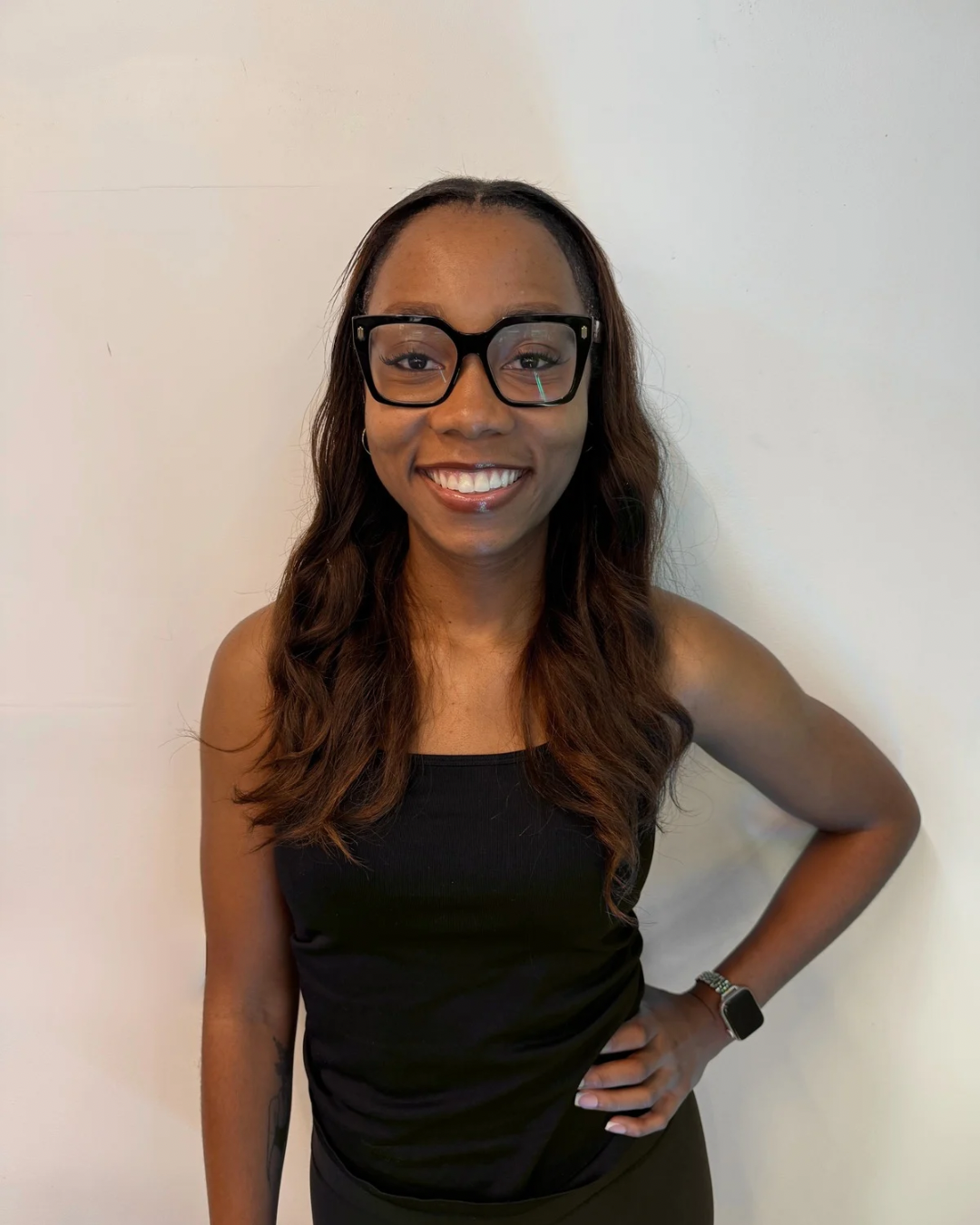 A woman with long wavy brown hair, wearing glasses, a black sleeveless top, and a smartwatch, standing against a plain white wall, smiling with her hand on her hip.