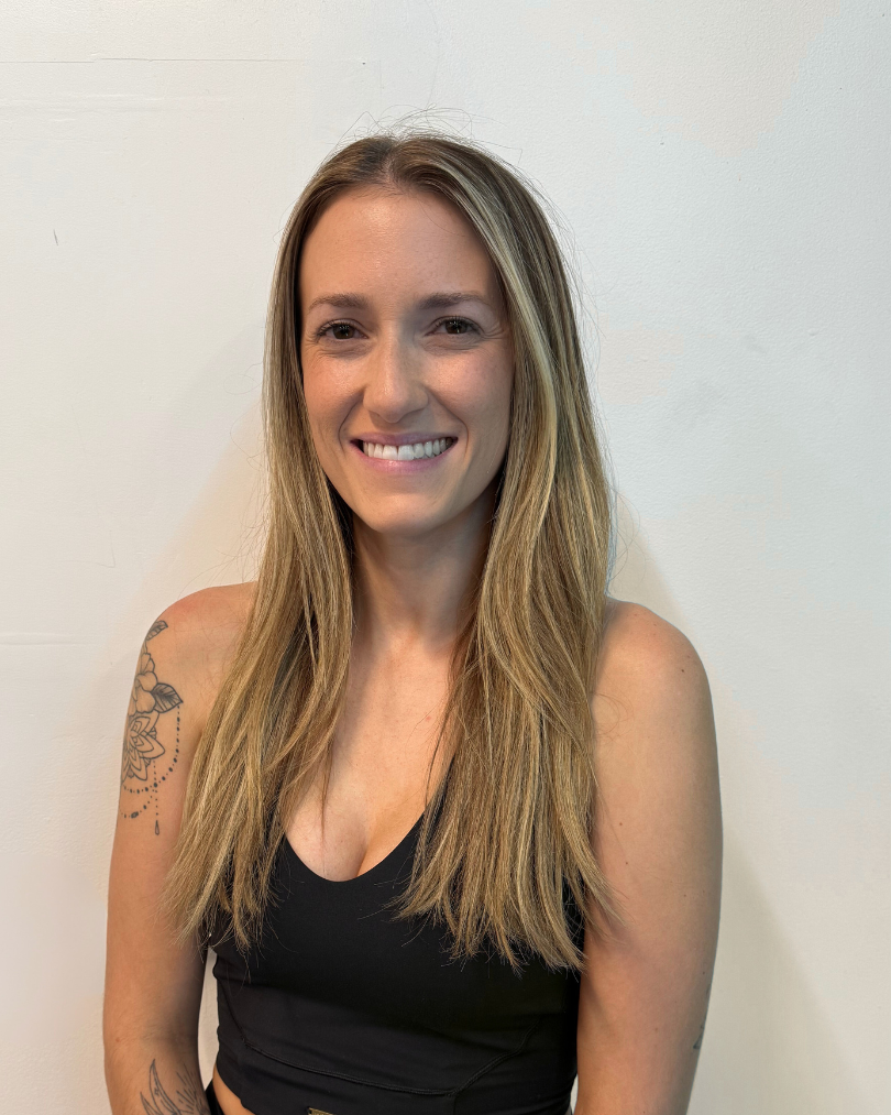 A smiling woman with long blonde hair, wearing a black tank top, standing against a plain white wall.