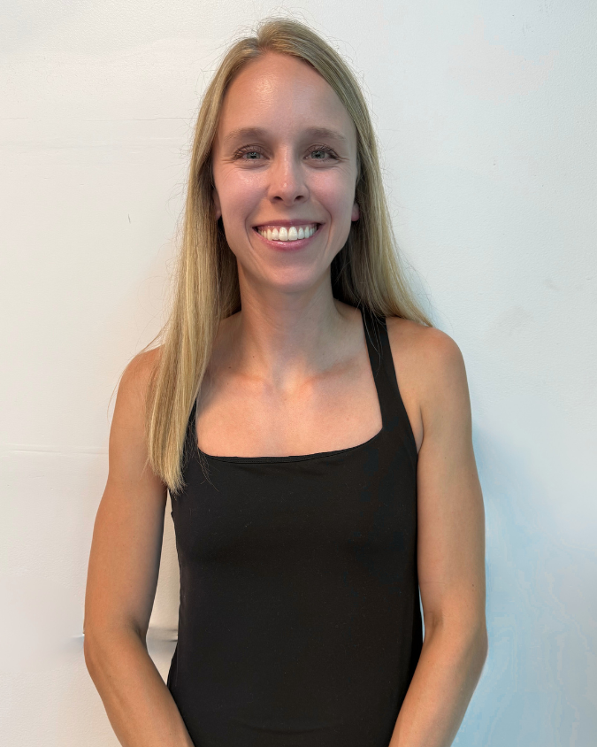 Smiling woman with long blonde hair wearing a black sleeveless top standing against a white wall.