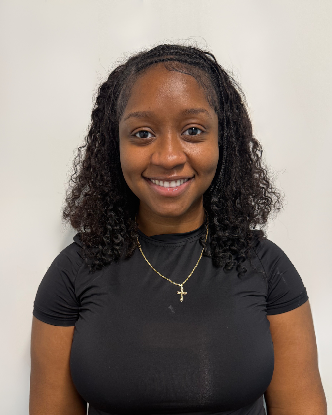 A smiling young woman with dark curly hair wearing a black athletic shirt and a gold cross necklace, standing against a plain white background.