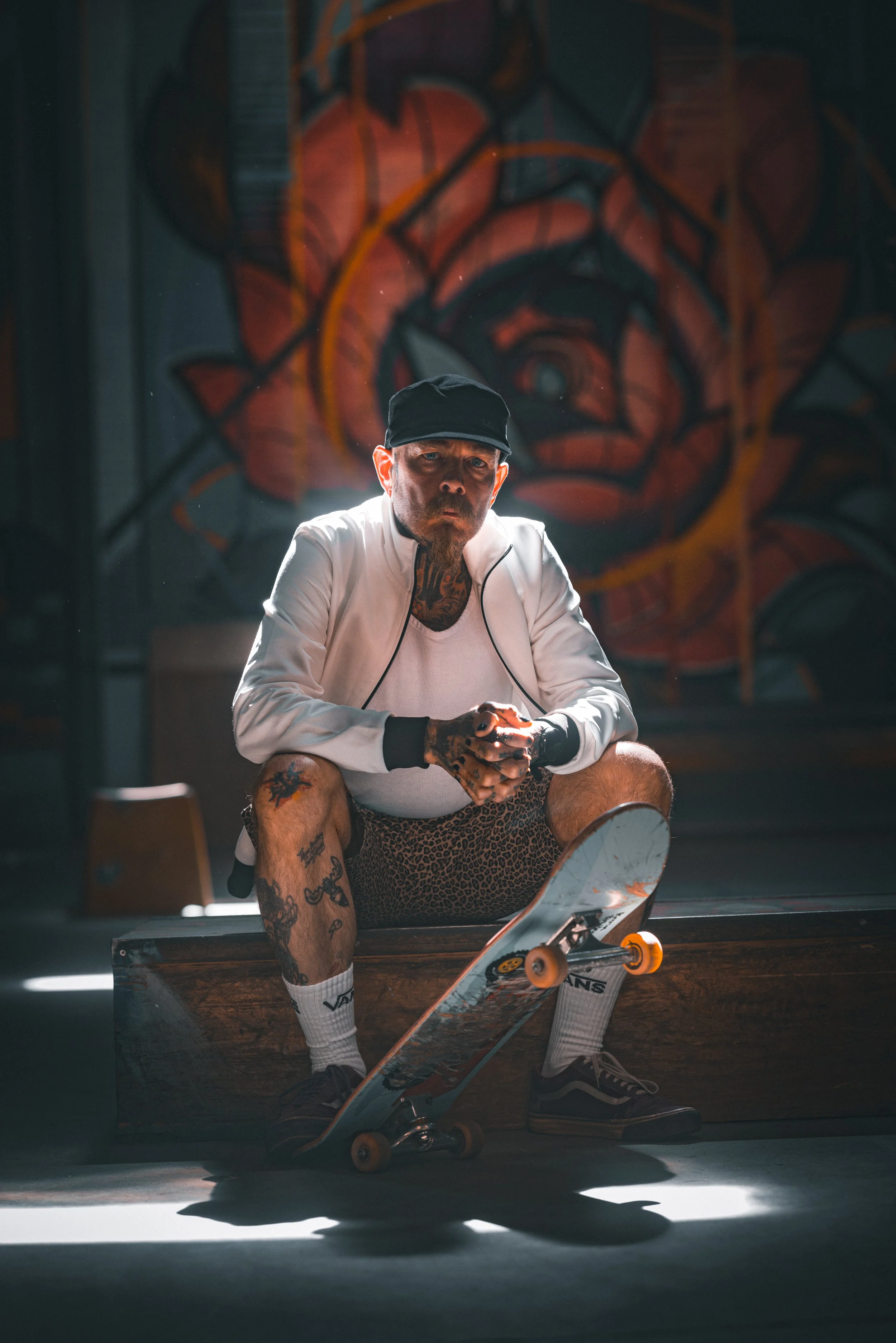 A tattooed man with a beard, wearing a black cap, white jacket, and leopard-print shorts, sitting on a wooden ledge with a skateboard in a dimly lit skate park with graffiti art on the wall behind him.