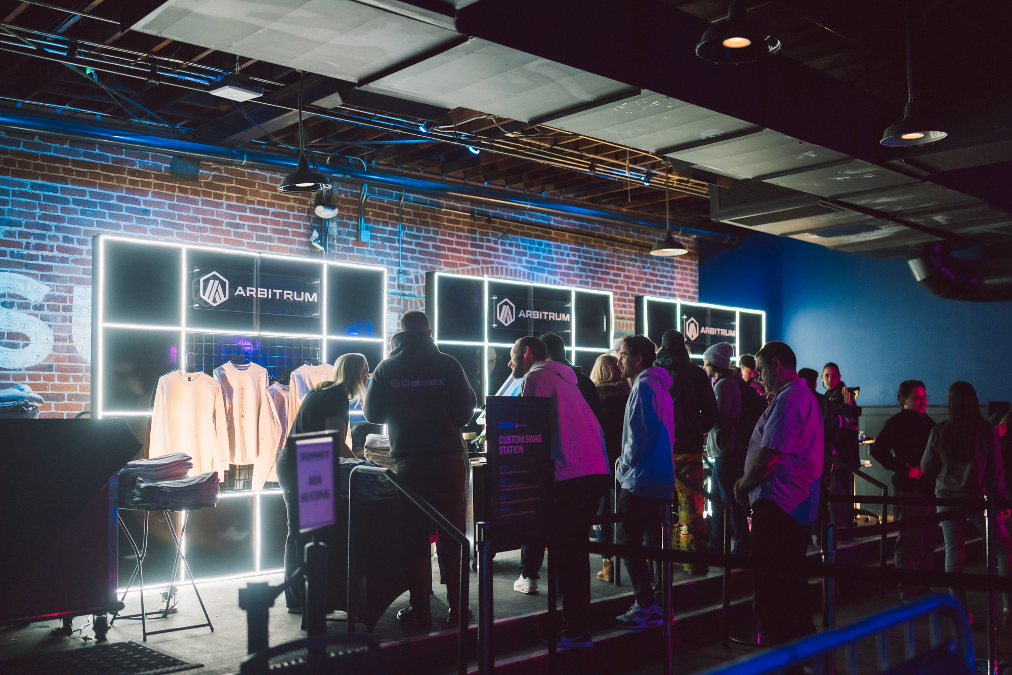 Line of people at a merchandise booth with illuminated signs that say 'Arbitrum' inside a dimly lit venue.