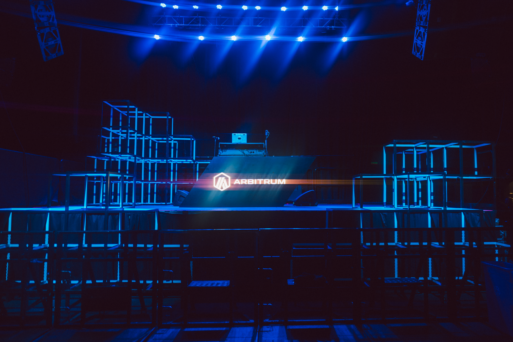Empty stage with blue lighting, Arbitrum logo on a large screen, and geometric metal framework structures on either side, in a dark indoor setting.