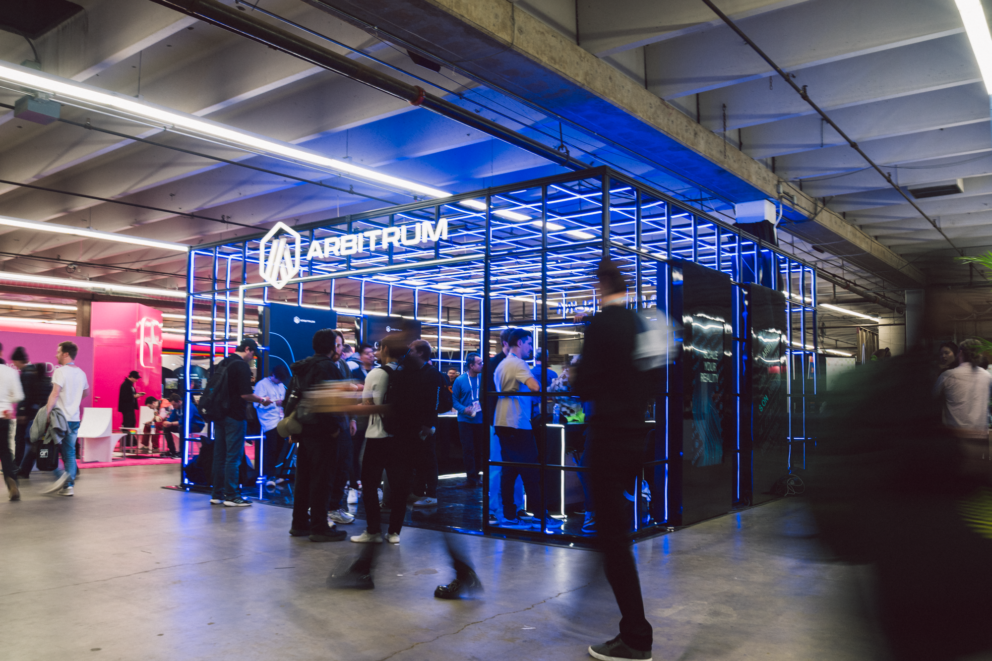 A large indoor exhibition booth with the sign 'Arbitrum' made of neon lights, surrounded by a crowd of people, in a spacious industrial-style venue.