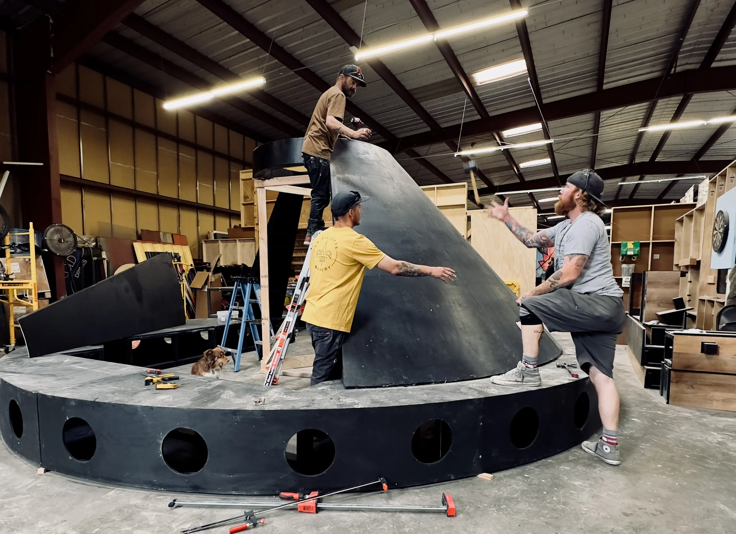 Four men working on a large, black, cone-shaped structure inside a workshop with wooden shelves and various tools. A small dog is sitting on the edge of the structure. One man is standing on a ladder, another is gesturing as if giving instructions, and two others are standing nearby engaged in conversation.