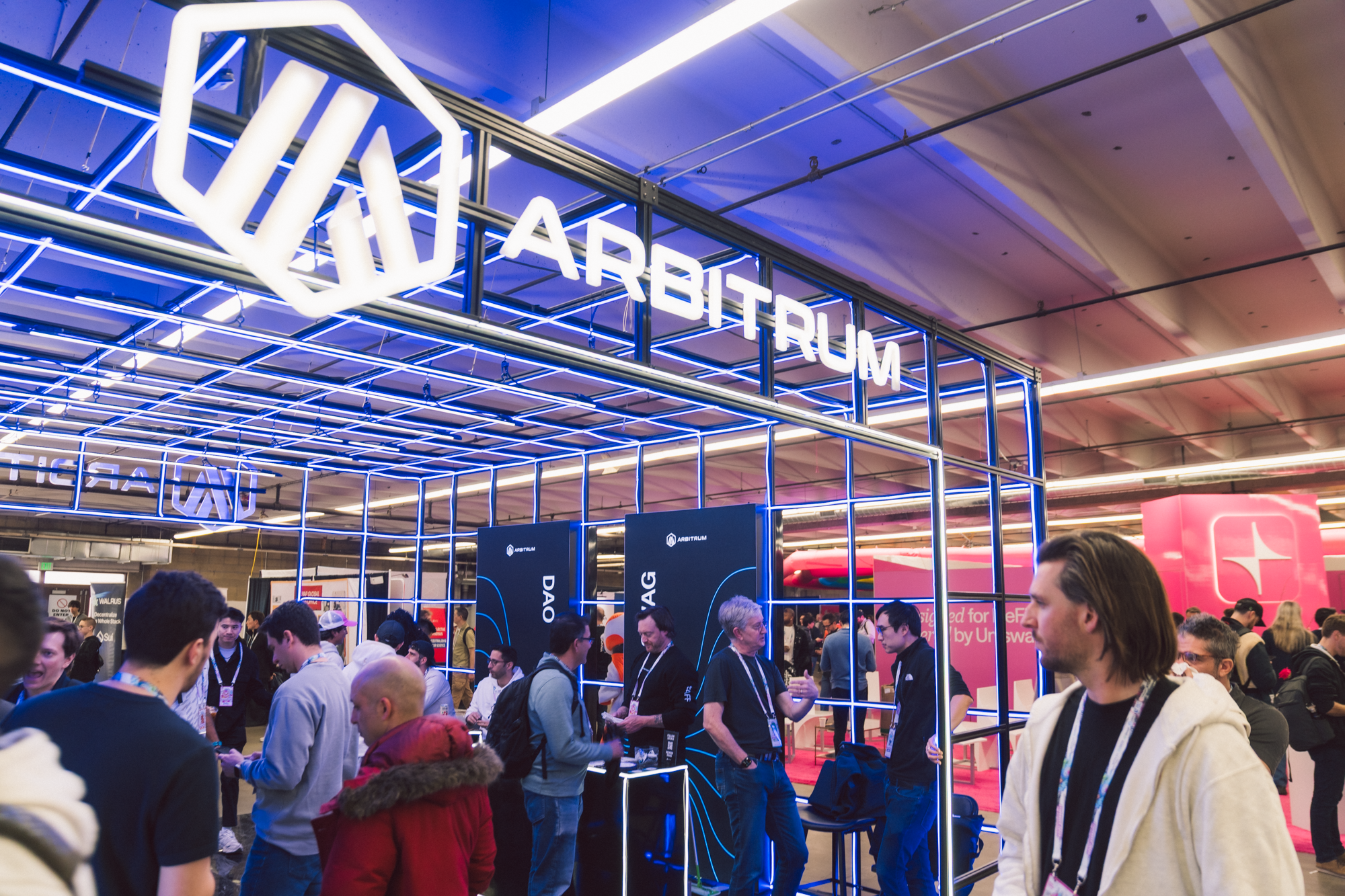People gathered at the ARBITRUM booth during a conference, with a prominent neon sign displaying 'ARBITRUM' and a geometric logo, inside a large indoor event space.
