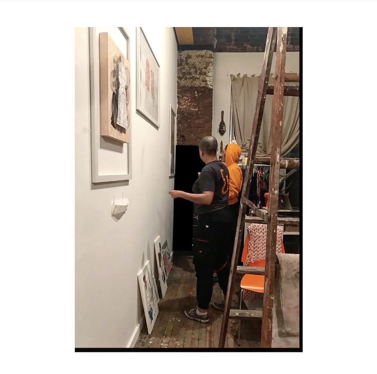 Two people stand in an art gallery or studio, looking at framed artwork on a white wall. One person wears a black shirt with an orange design, and the other wears an orange hoodie. There is a wooden stepladder leaning against the wall to the right, a