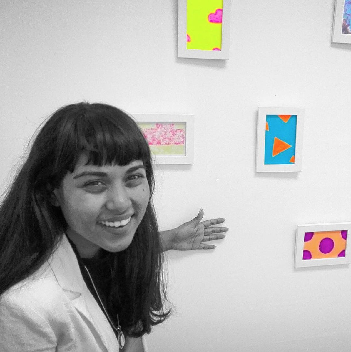 A smiling woman with long dark hair pointing at colorful, abstract artwork on a white wall in an art gallery.