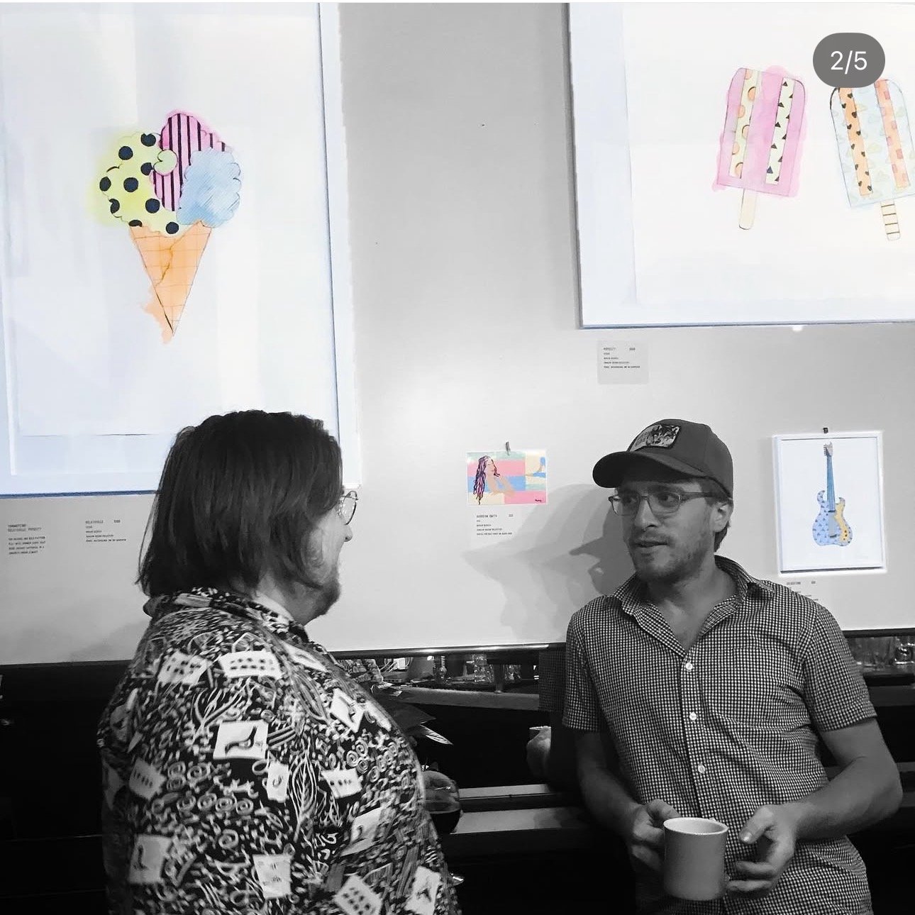 Two men are talking in an art gallery; one is holding a cup. There are colorful art pieces, including an ice cream cone and popsicles, hanging on the wall behind them.