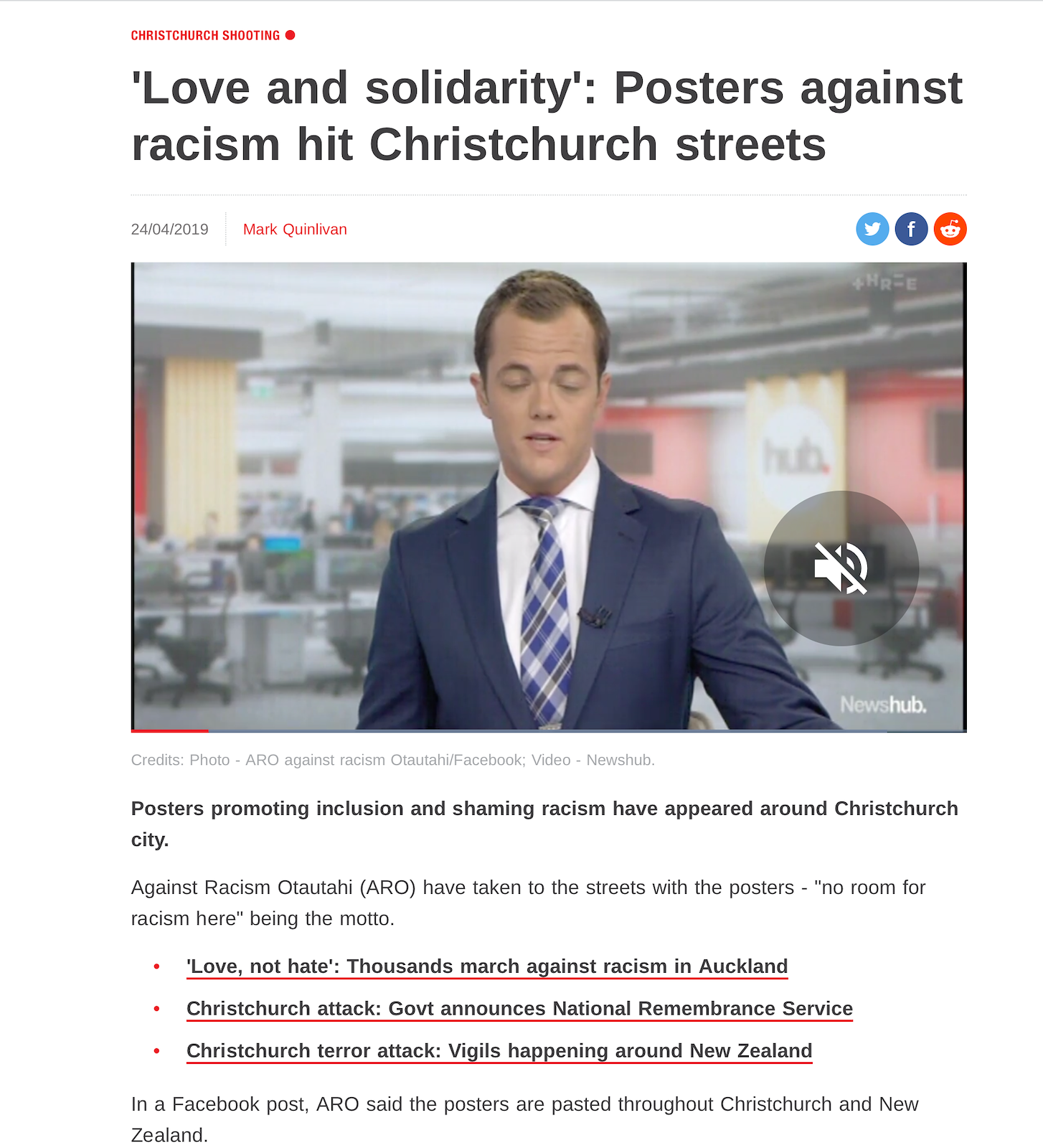 A news reporter in a newsroom with a blurred background featuring various screens and office furniture. He is wearing a dark blue suit, white shirt, and diagonal striped tie, and appears to be reading or speaking.