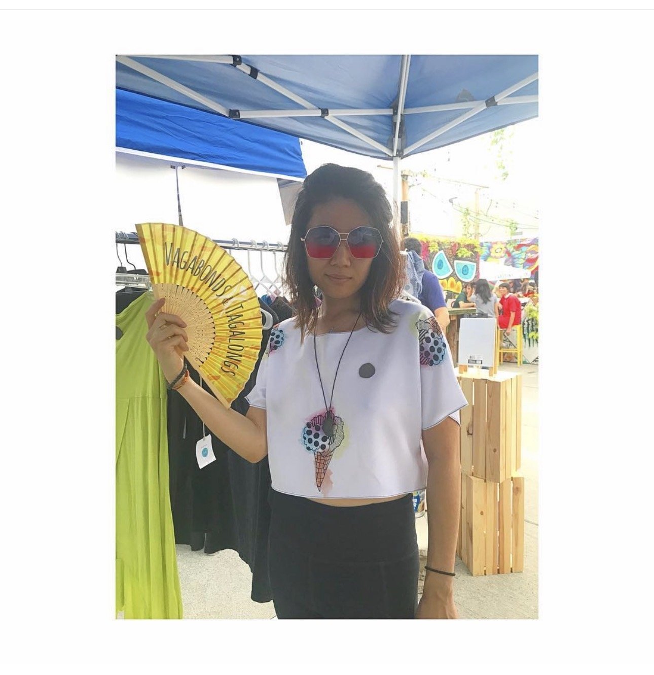 A woman wearing sunglasses holding a yellow fan with the words "VAGABOND VAGALONGS" written on it. She is at an outdoor event under a canopy, with colorful decorations and other people in the background.