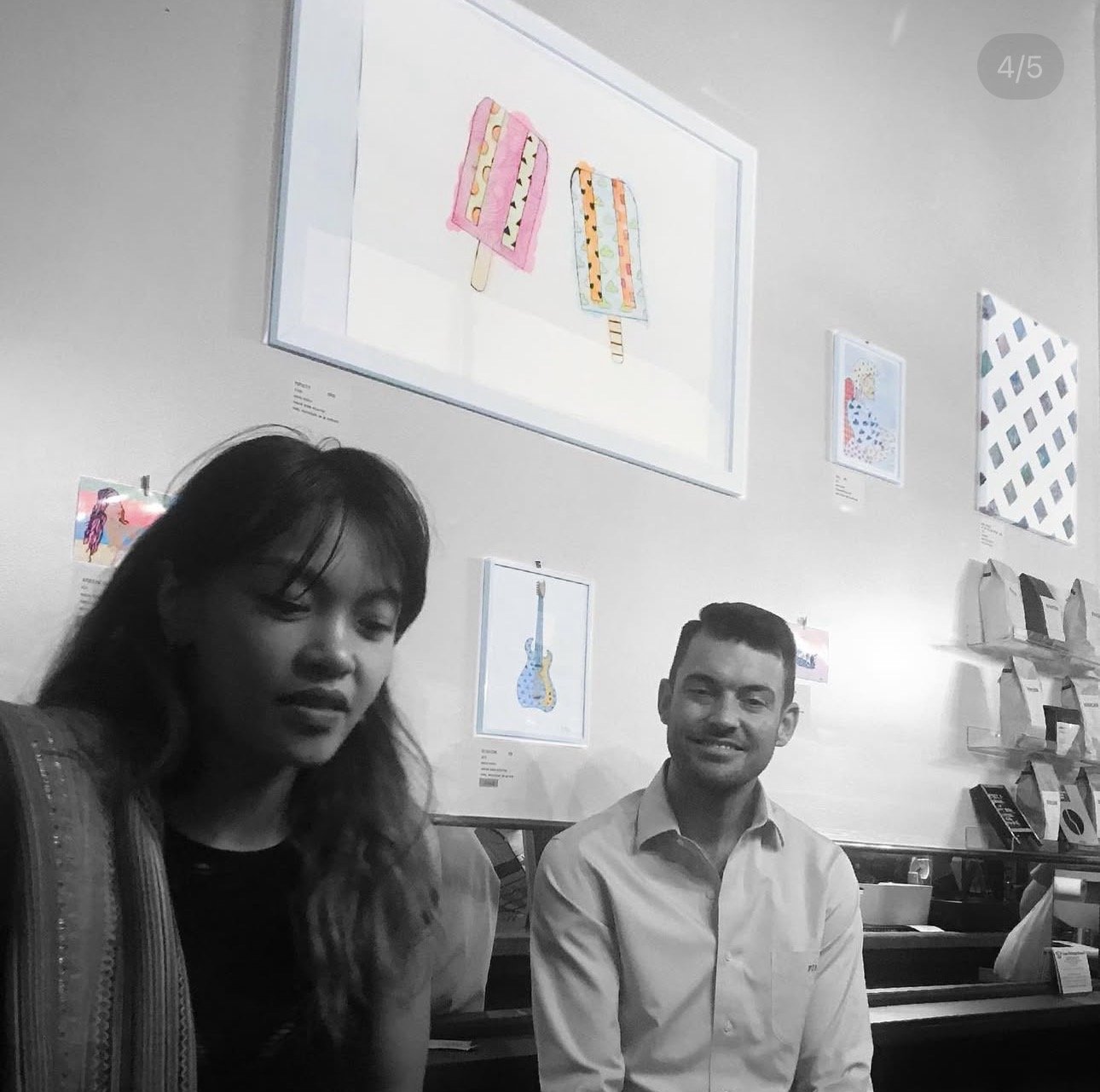 A woman and a man sitting and talking in an art gallery with framed illustrations, including colorful ice cream popsicles and guitars, displayed on the white wall behind them.