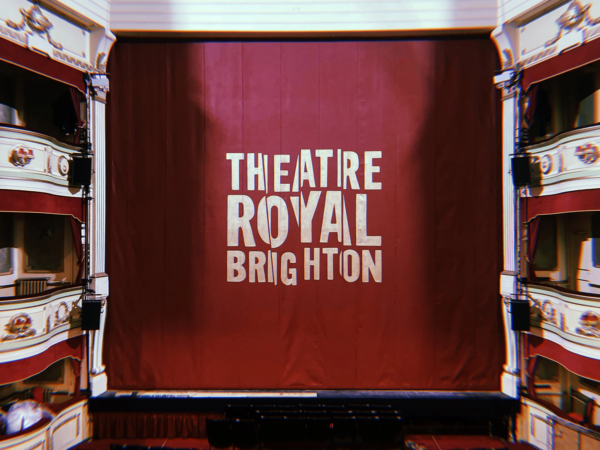 The interior of a theater showing a large red curtain with the logo for 'Theatre Royal Brighton' written on it in large white letters.