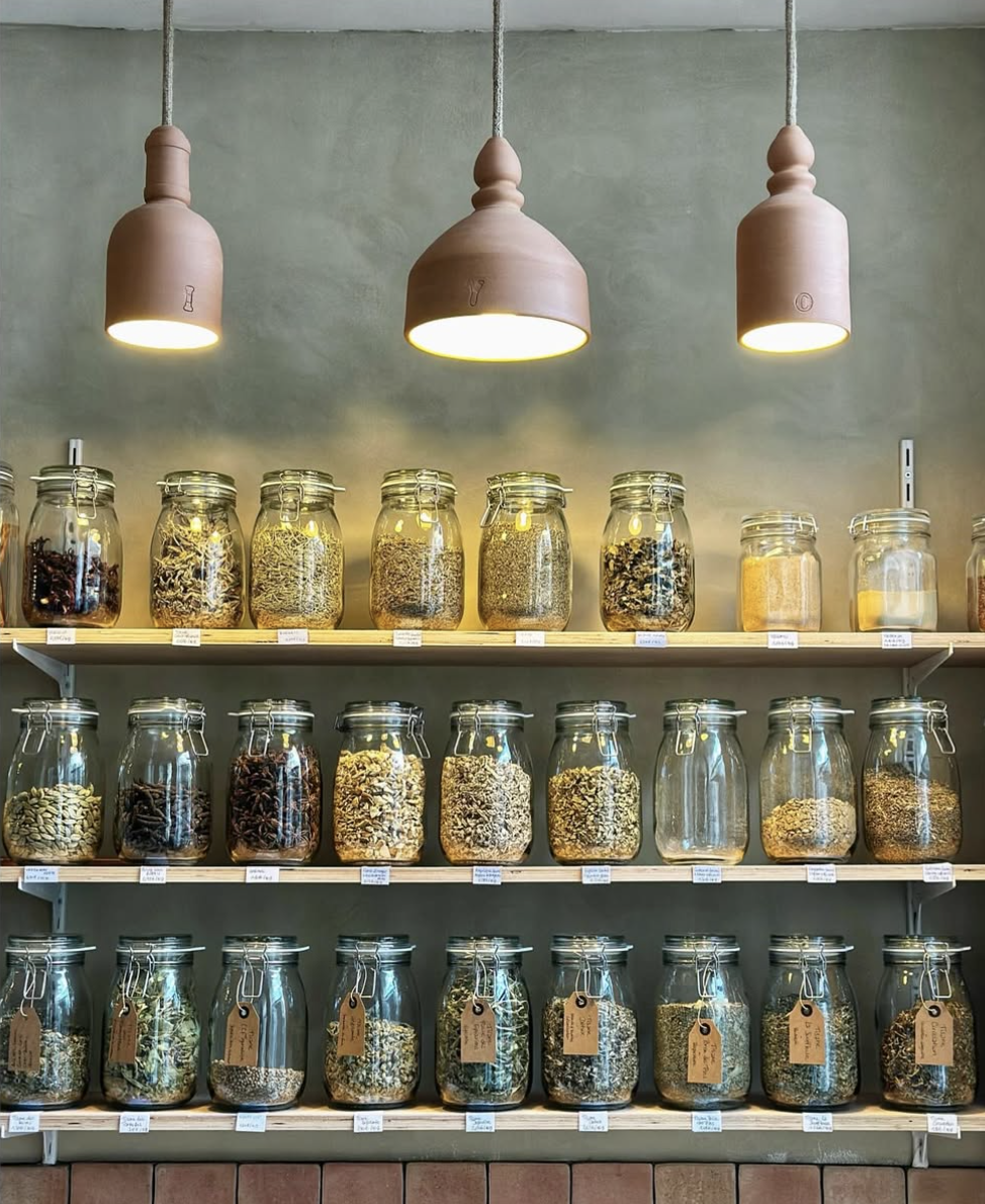 Étagère avec des bocaux en verre contenant des épices ou des herbes, éclairés par des lampes suspendues au plafond.