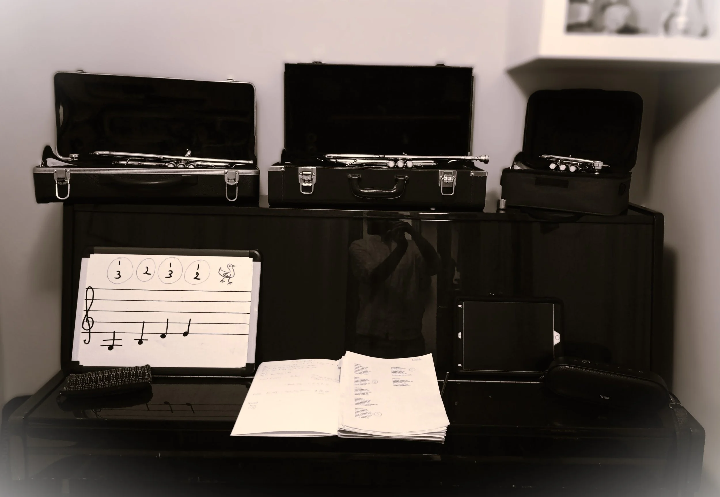B&W photo of three trumpets in open cases resting on top of an upright piano. A music score and speaker are visible on the closed keyboard lid.