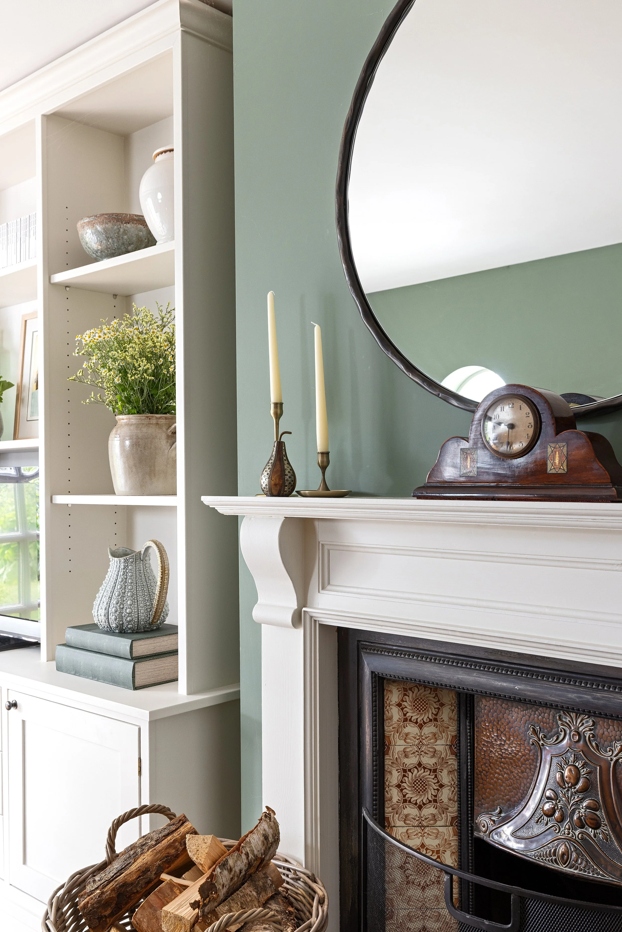 A living room corner with a white fireplace, a large round mirror, and a wooden mantel, decorated with a vintage clock and candle holders. To the left of the fireplace is a white bookshelf with decorative vases, a potted plant, and books. In front of