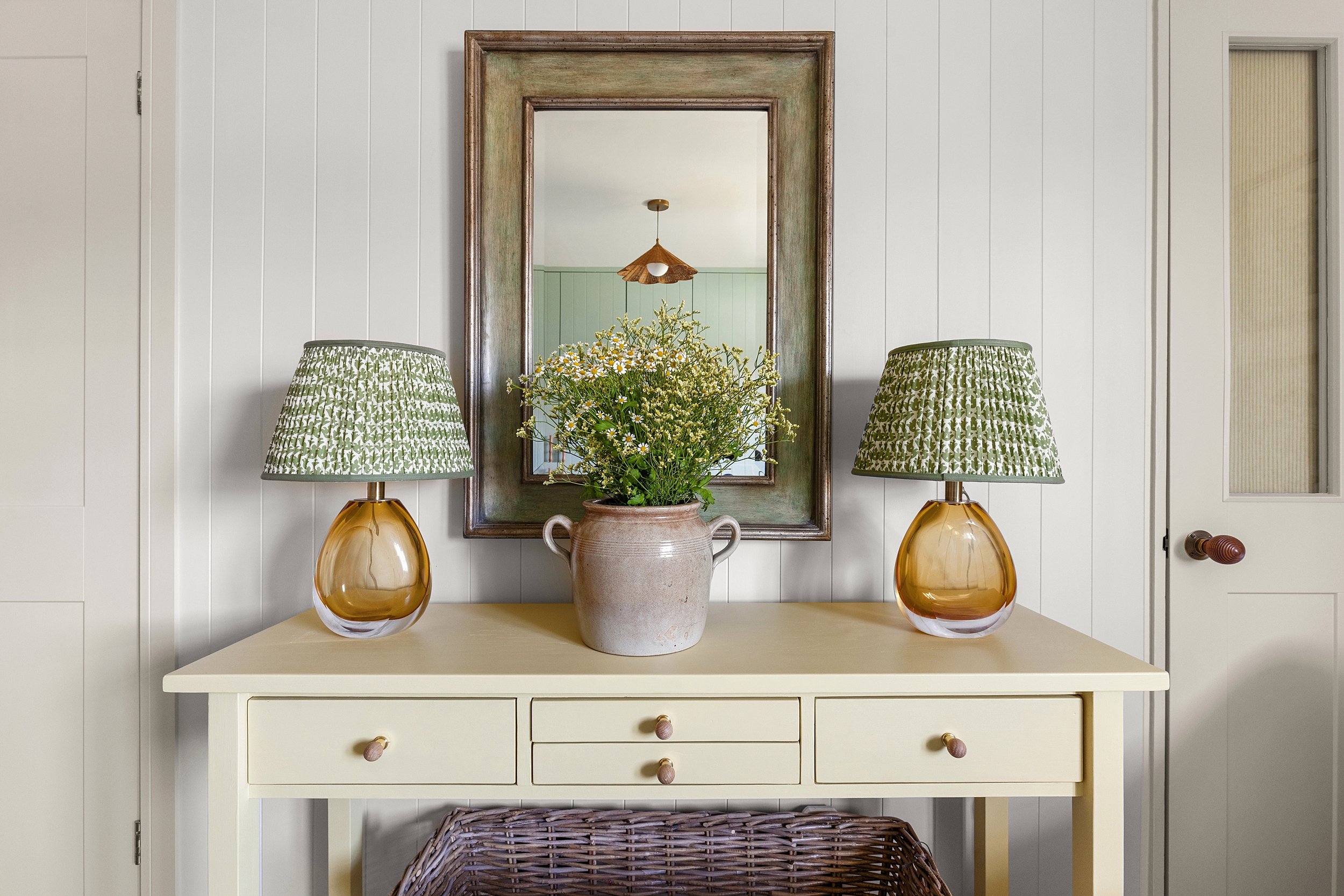 A cream-colored sideboard with three drawers, decorated with a potted plant and two matching lamps with green patterned shades on either side. A large framed mirror hangs above, reflecting a ceiling light and part of a room with green walls.