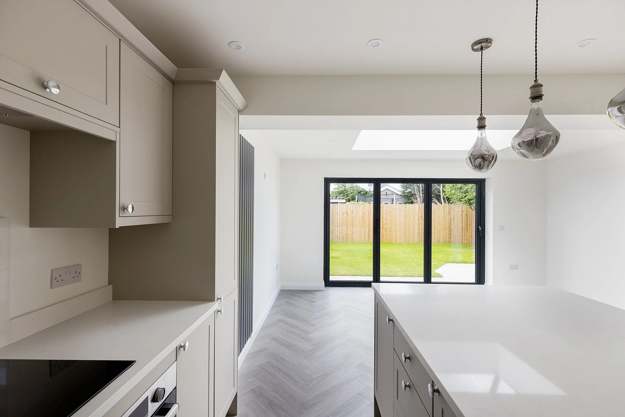Empty modern kitchen with white cabinets and countertops, hanging pendant lights, and large glass sliding doors opening to a backyard with grass and a wooden fence.