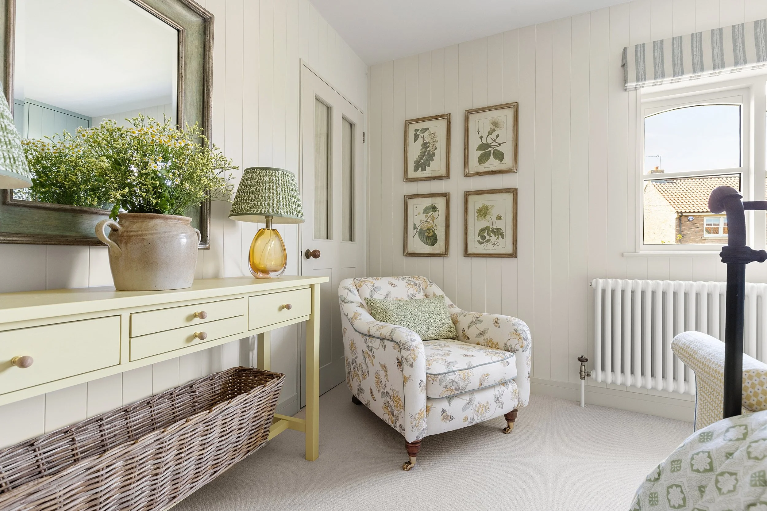 Interior of a cozy room with a cream-colored armchair with floral pattern, four botanical prints on the wall, a cream sideboard with a lamp and a vase of greenery, a mirror, and a window with a view of neighboring rooftops.