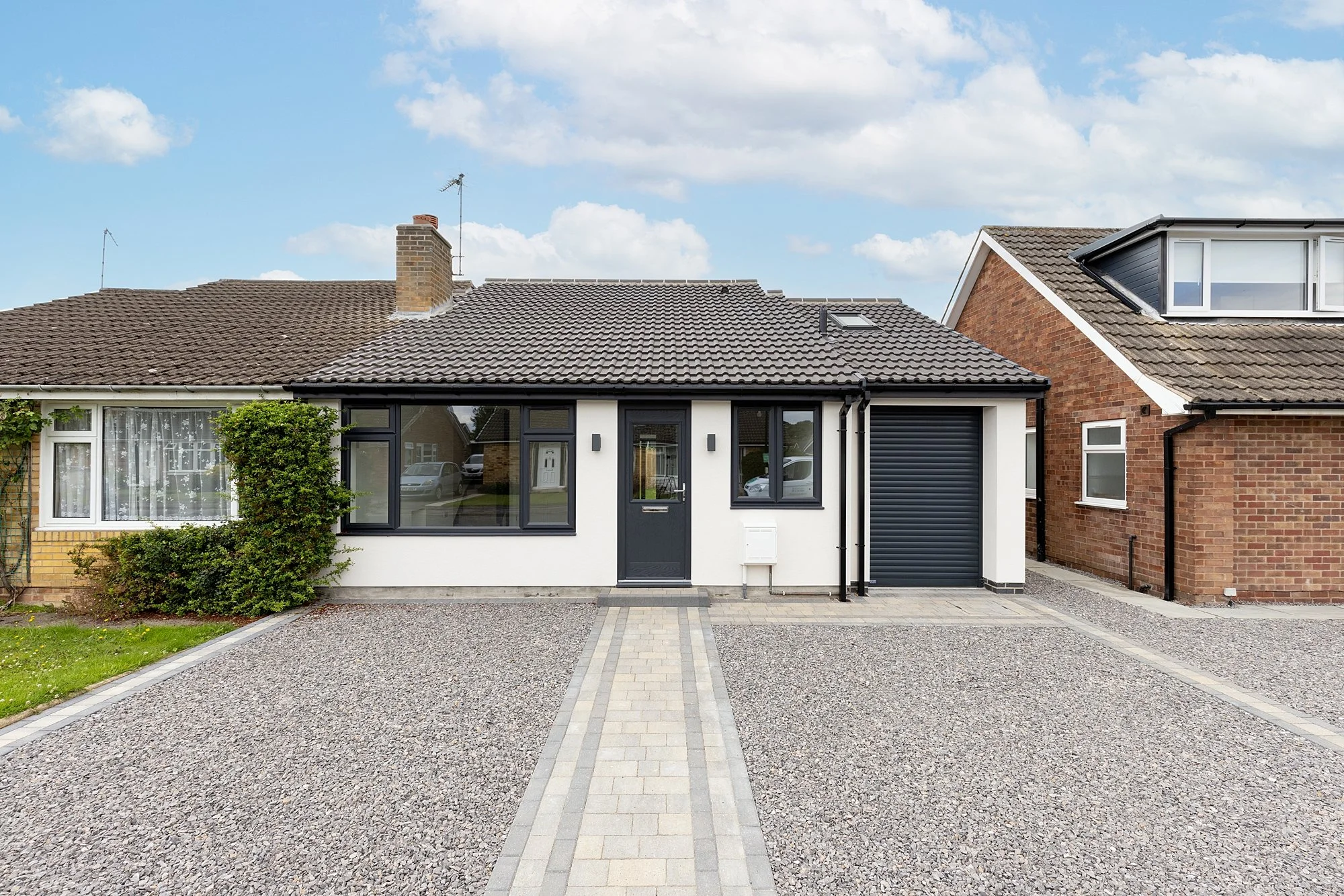 Front view of a modern house with a gravel driveway, white and black exterior, two windows, a front door, and an attached garage, under a partly cloudy sky.
