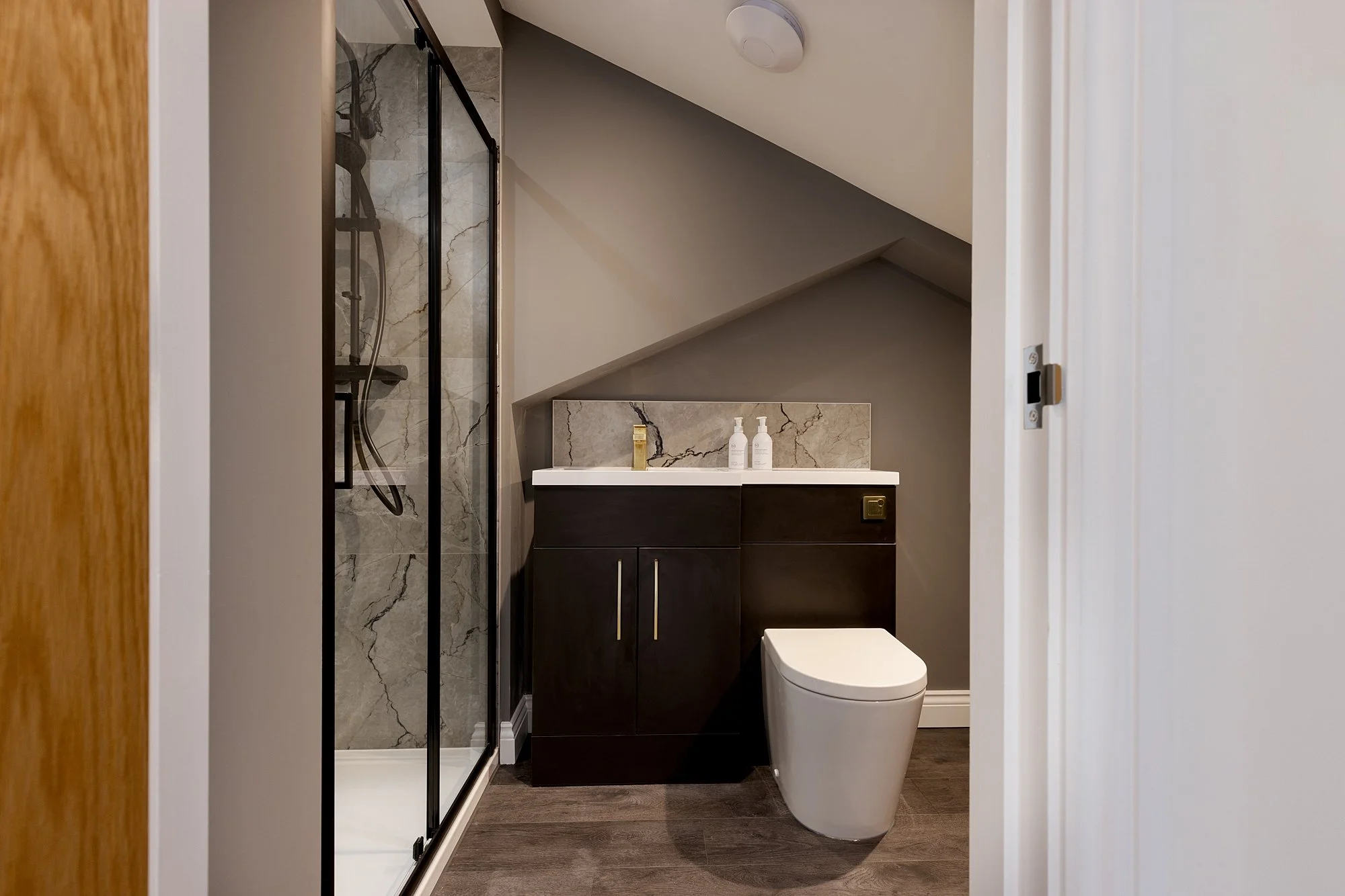 Small modern bathroom with a walk-in shower, black vanity, dual sinks, and a smart toilet, under a sloped ceiling.