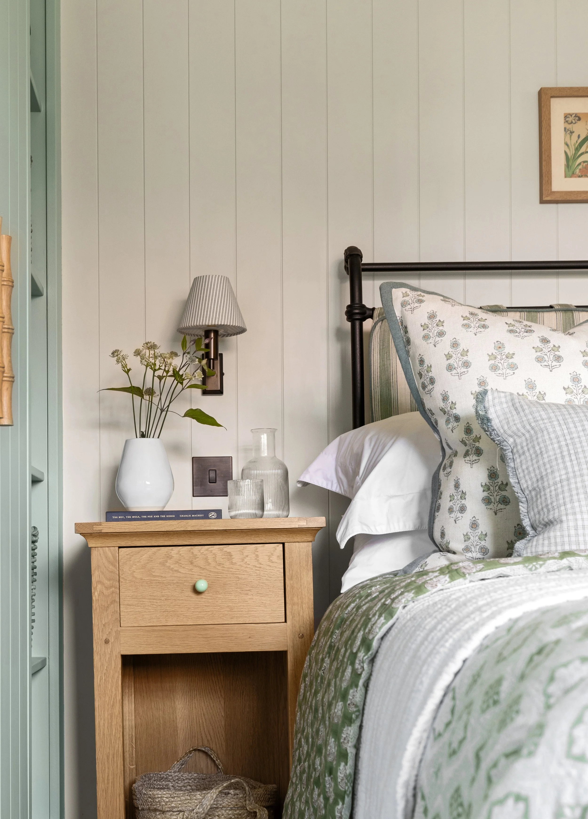 A bedroom scene showing a bed with floral and gingham pillows, a wooden nightstand with a glass vase with flowers, a glass jar, and a book, a wall-mounted lamp with a striped shade, and framed artwork on a white paneled wall.