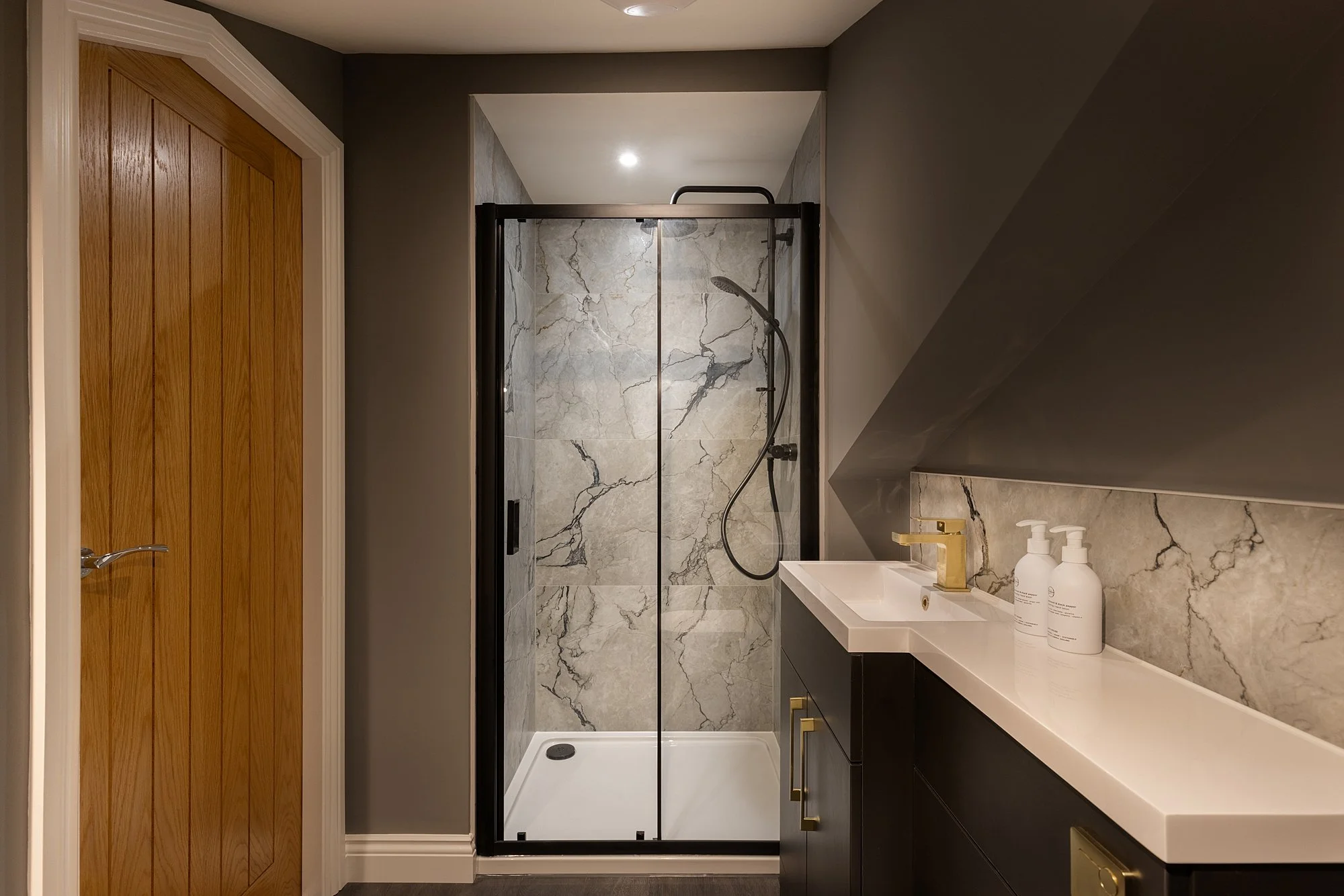 Modern bathroom featuring a walk-in shower with marble walls, a black framed glass door, a dark vanity with gold handles, a white countertop, and a gold faucet.